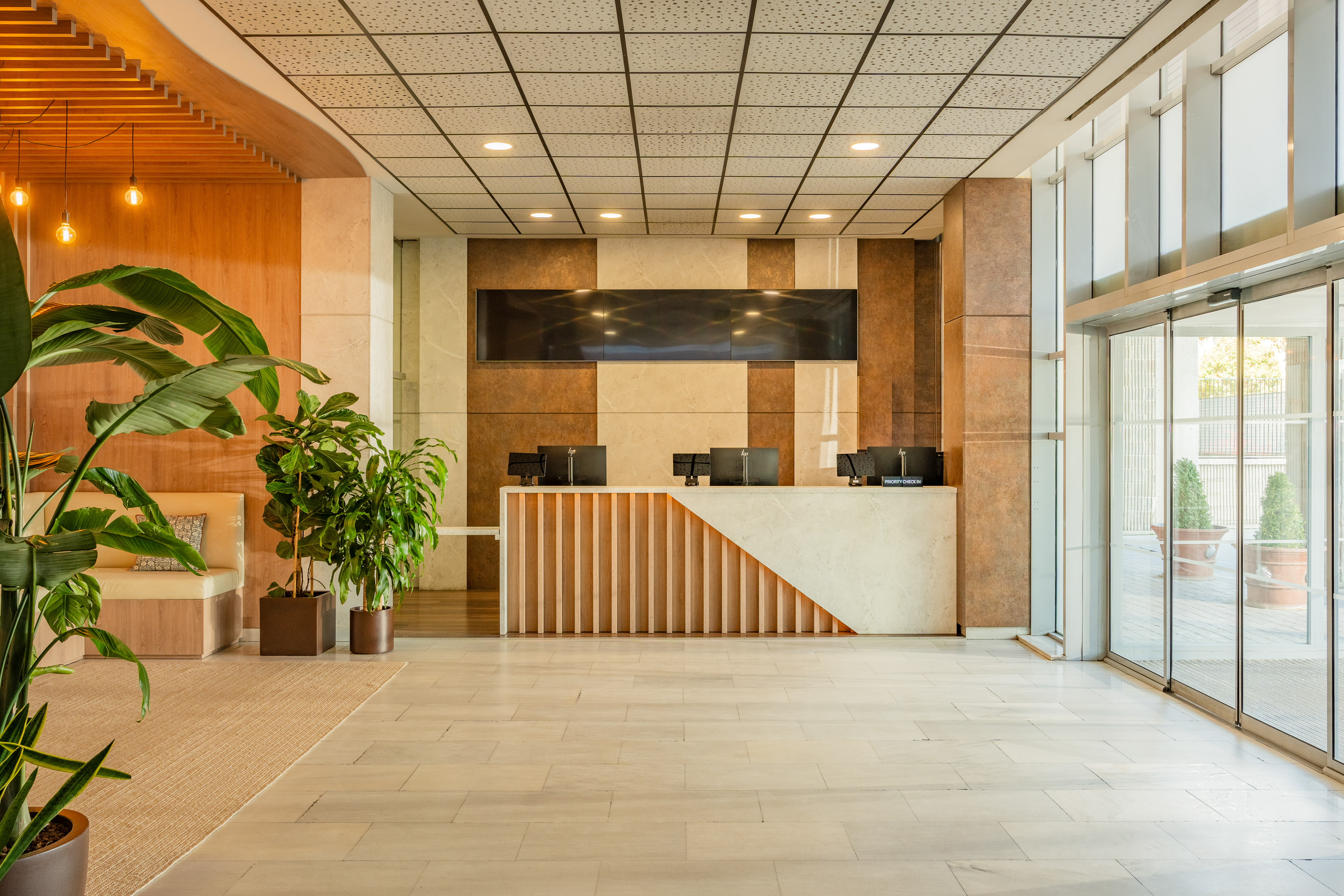 Modern hotel reception with wood and marble desk, green plants, glass doors.