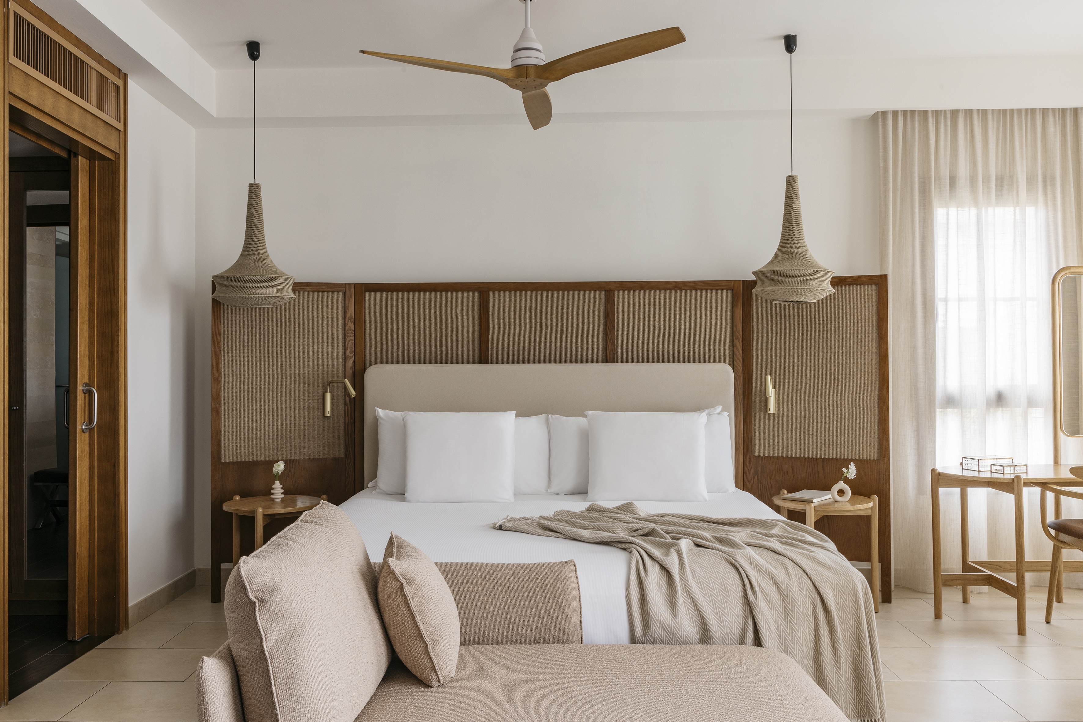 Modern hotel bedroom with king bed, cane headboard, wooden fan, woven lights.