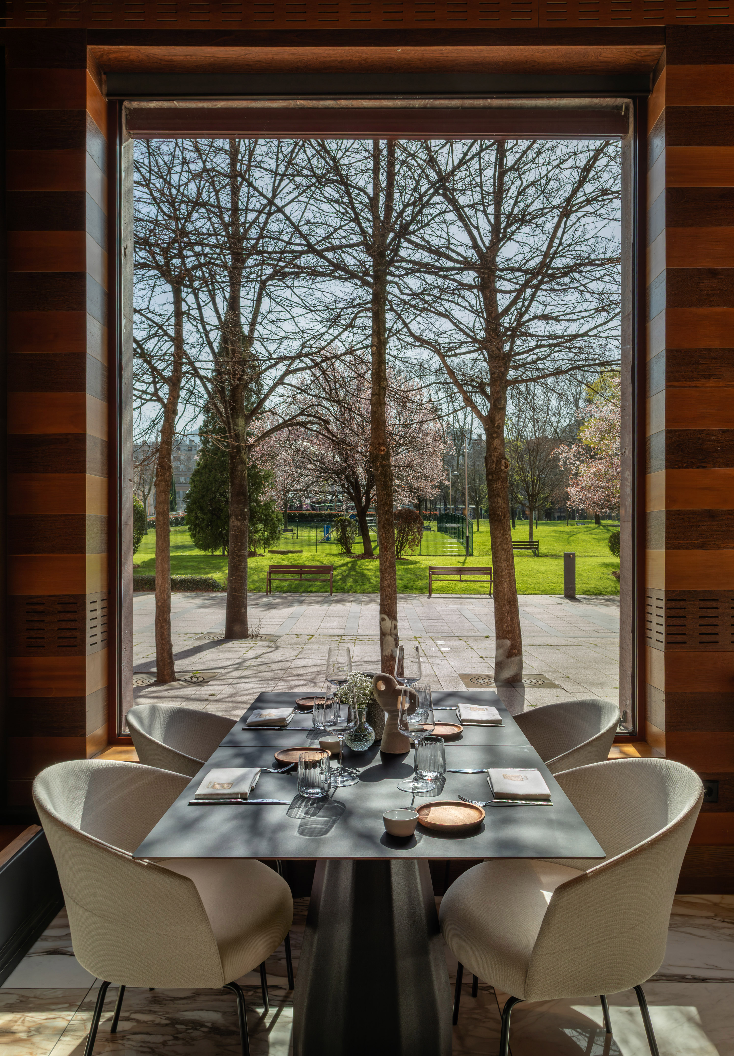Modern restaurant dining table by window, overlooking park with trees.