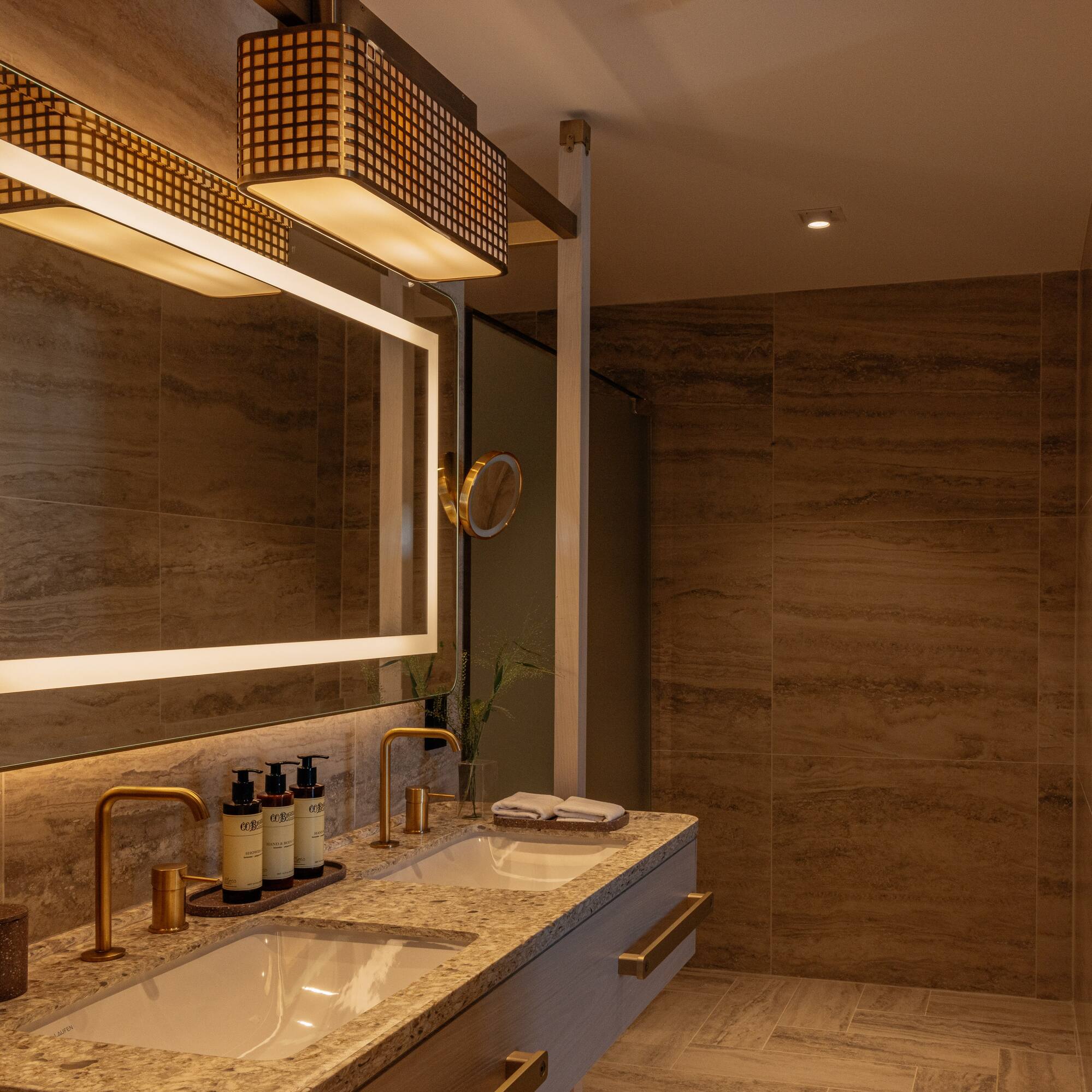 Modern bathroom with dual sinks, backlit mirror, gold faucets, and tiled walls.