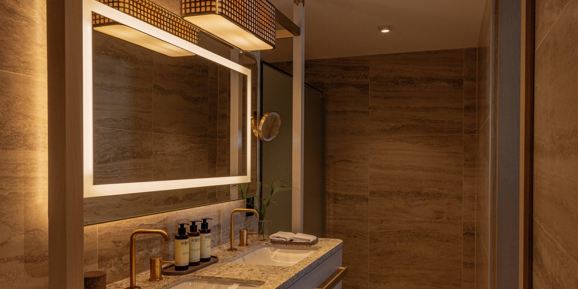 Modern bathroom with dual sinks, backlit mirror, gold faucets, and tiled walls.