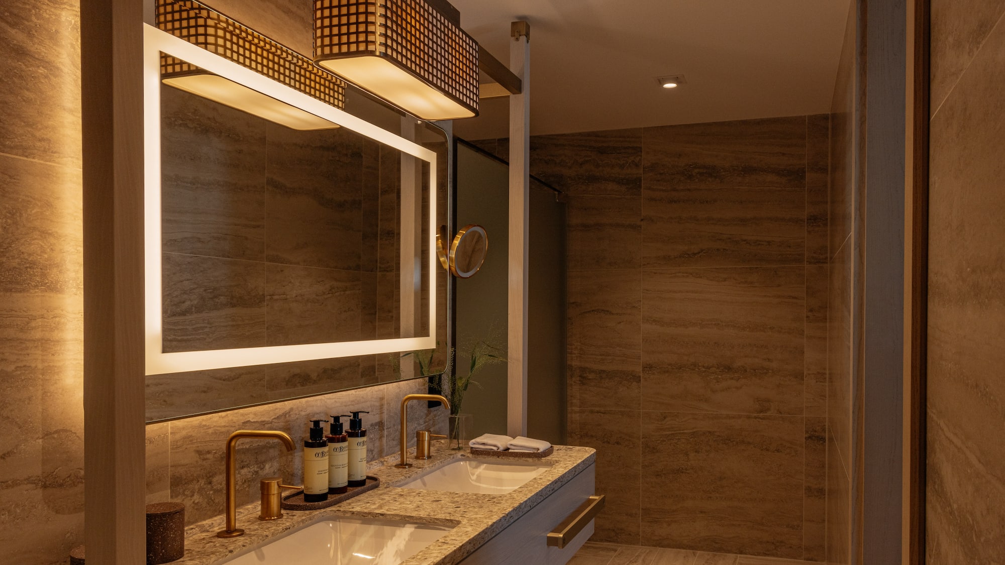 Modern bathroom with dual sinks, backlit mirror, gold faucets, and tiled walls.