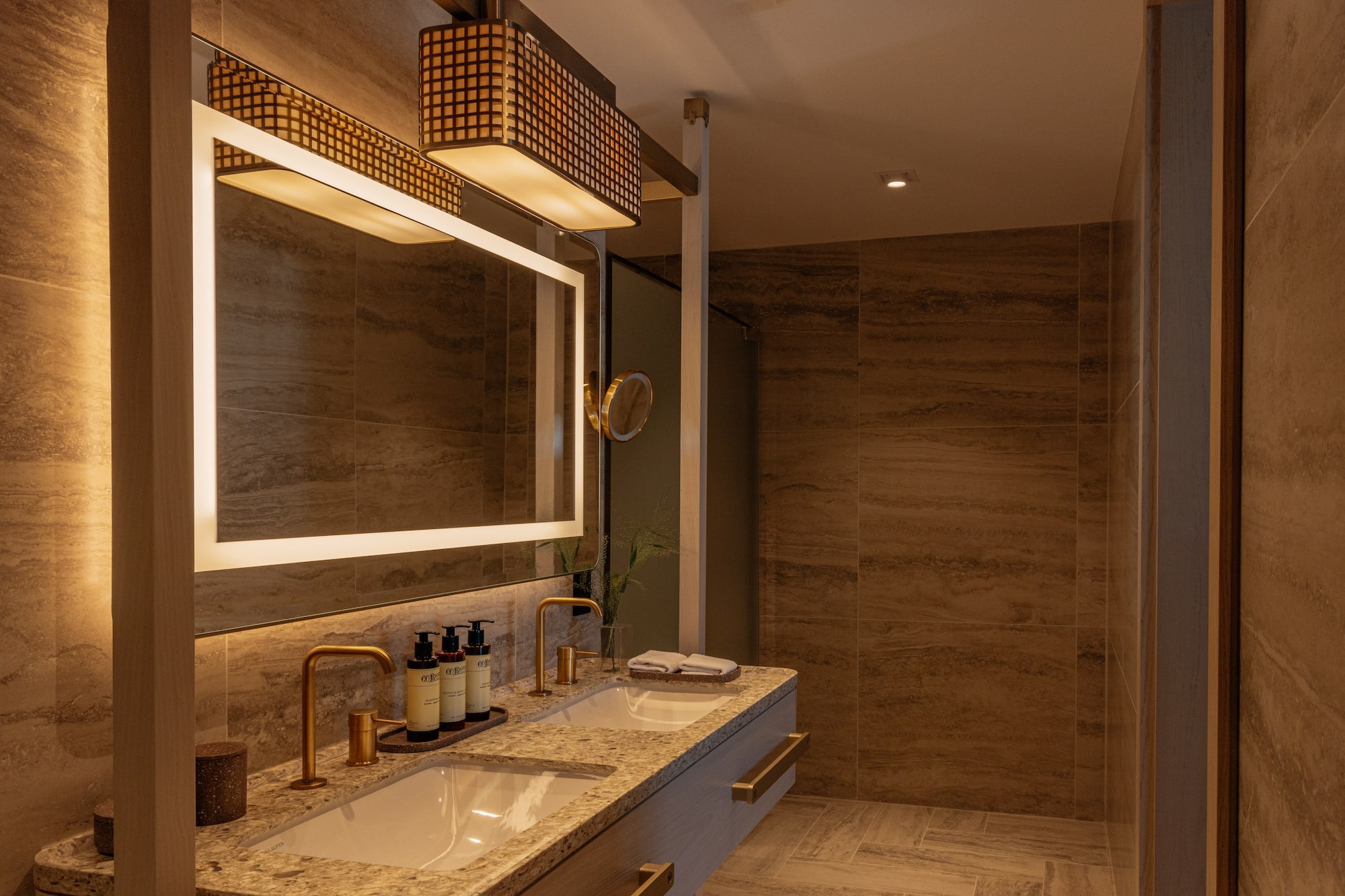 Modern bathroom with dual sinks, backlit mirror, gold faucets, and tiled walls.