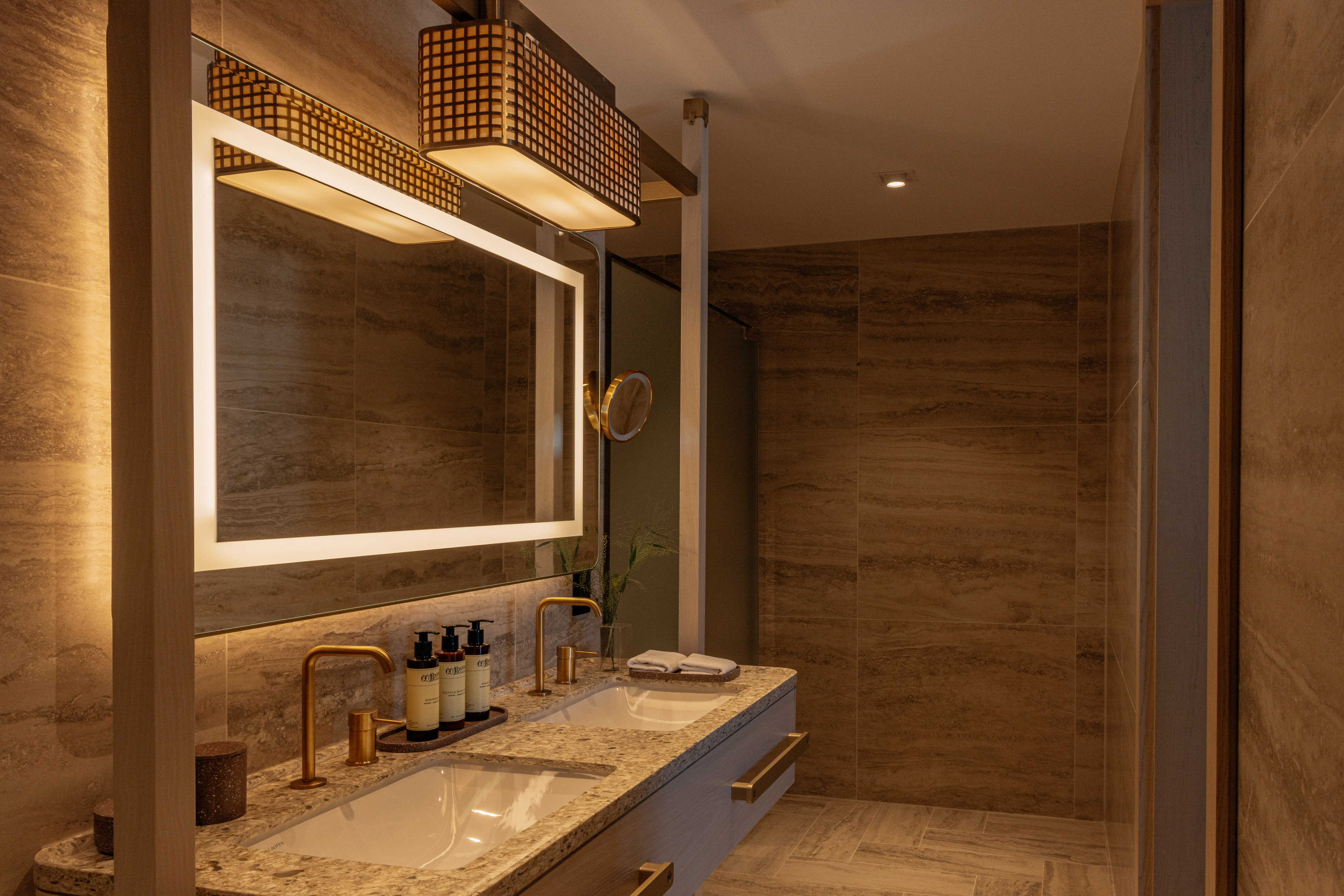 Modern bathroom with dual sinks, backlit mirror, gold faucets, and tiled walls.