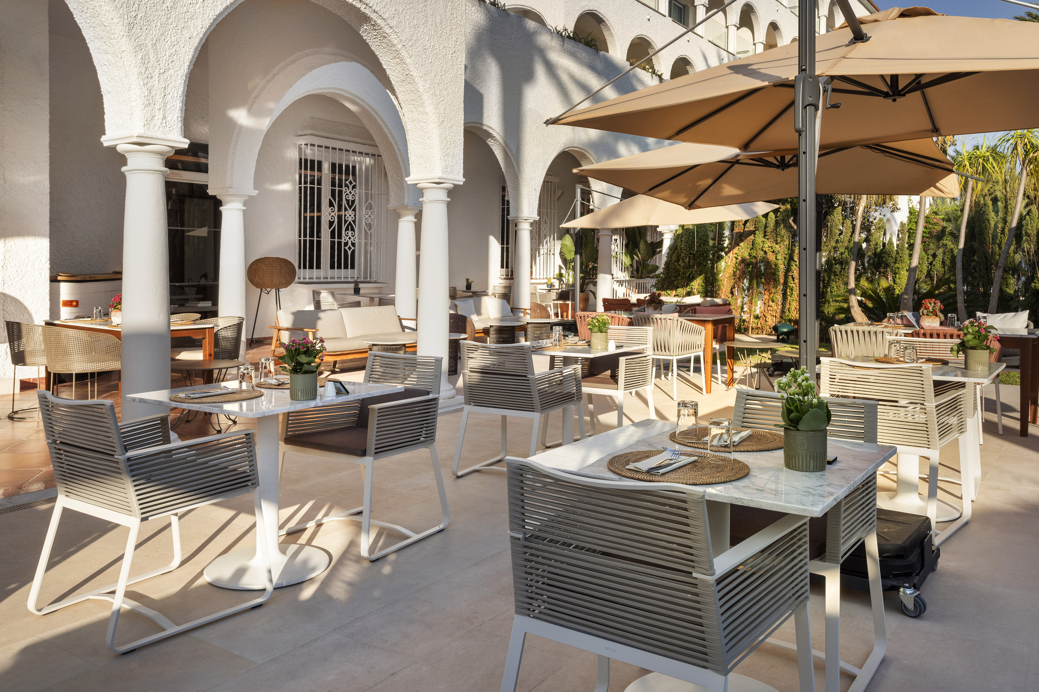 Outdoor patio restaurant with white arches, shaded tables, and lush garden.