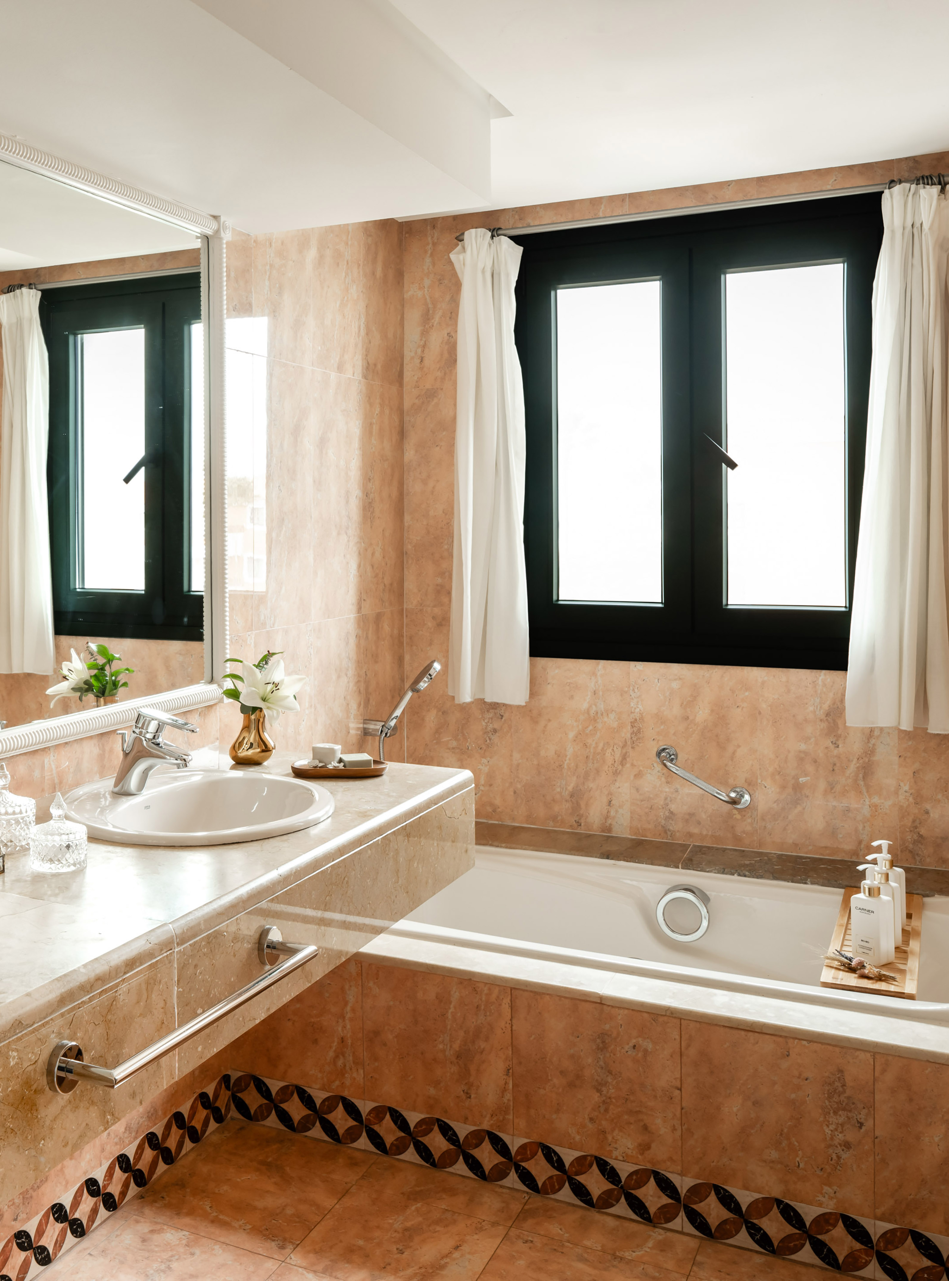 Modern bathroom with beige marble tiles, white sink, tub, and black windows.