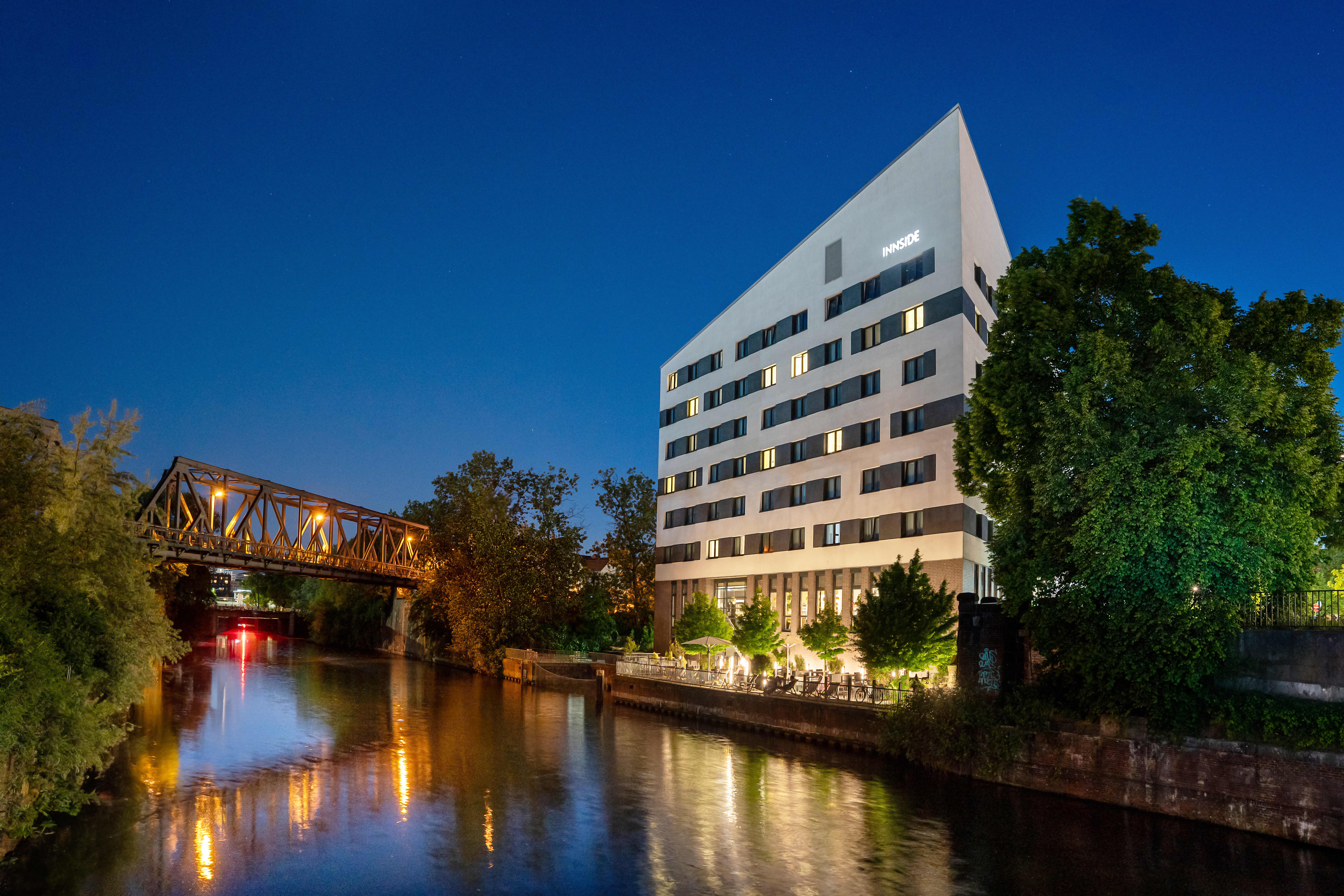 Modern INNSiDE Hotel at night, river, illuminated terrace, and bridge.