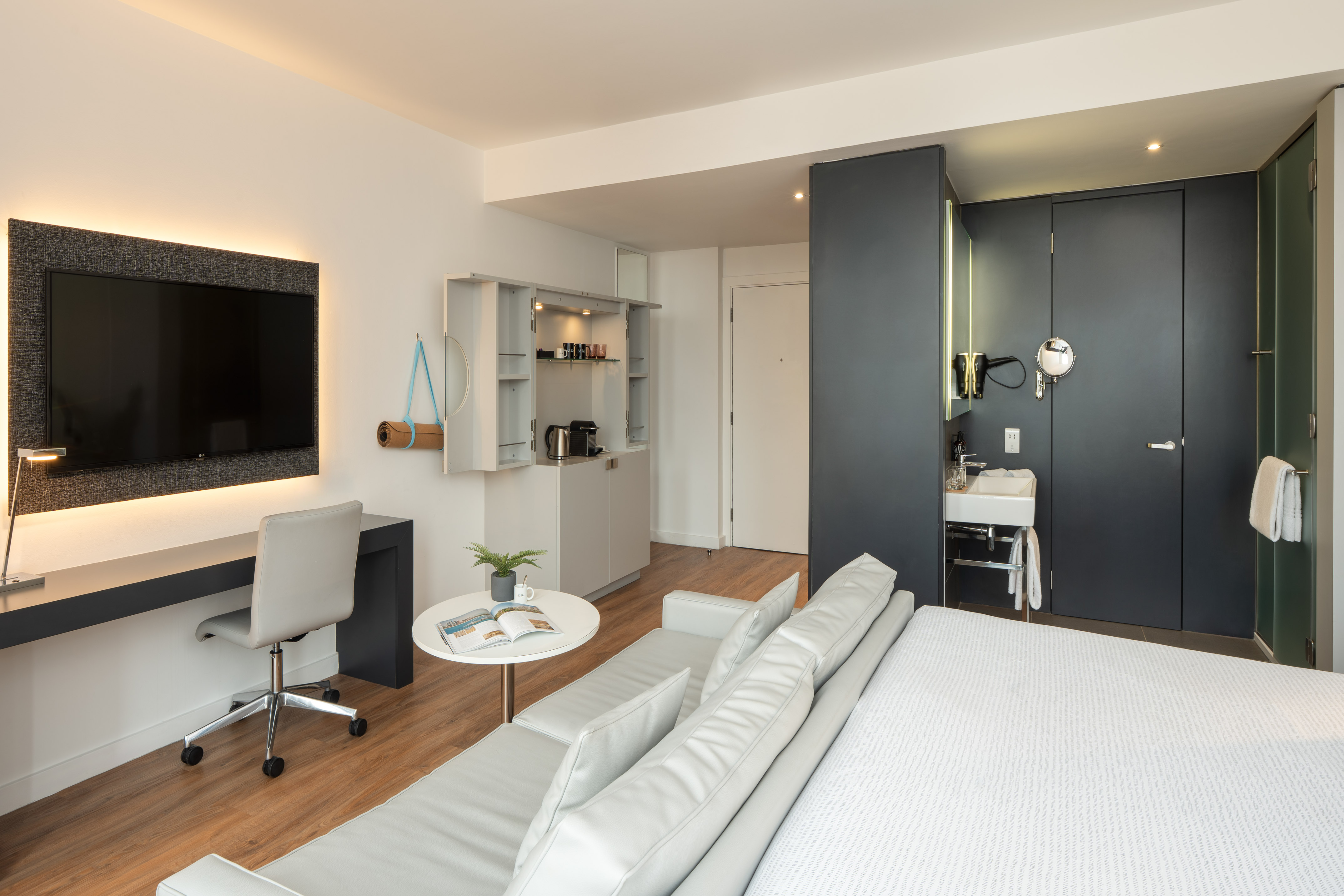 Modern hotel room with workspace, sofa, flat-screen TV, and integrated bathroom.