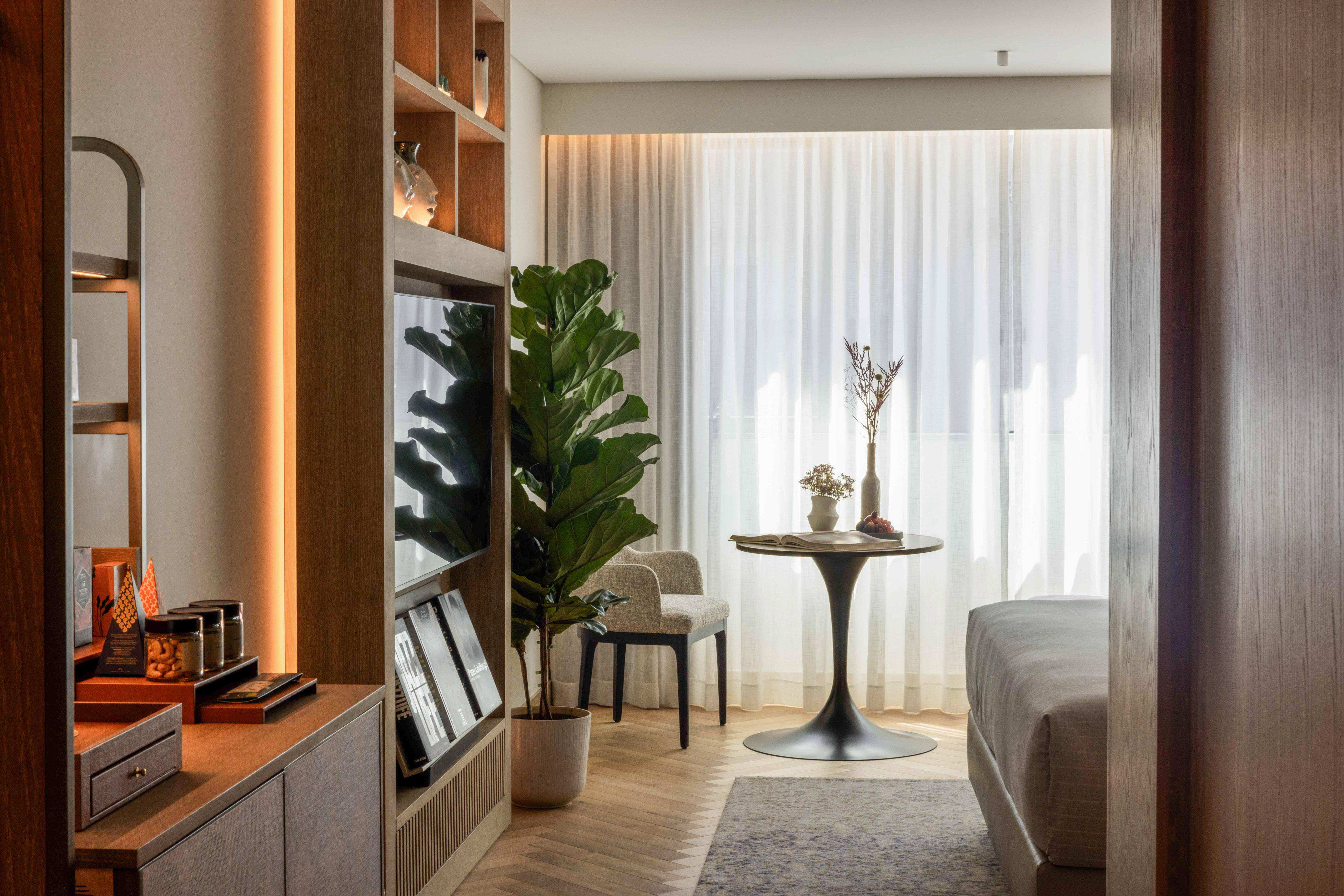 Modern hotel room with TV, dining area, large plant, and bright window.