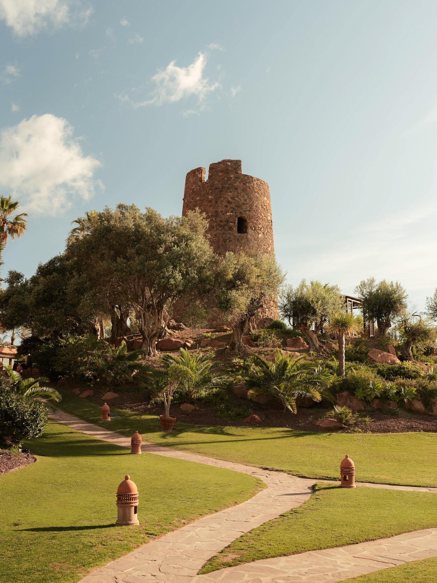 Old stone tower, winding path through a green garden with olive trees.