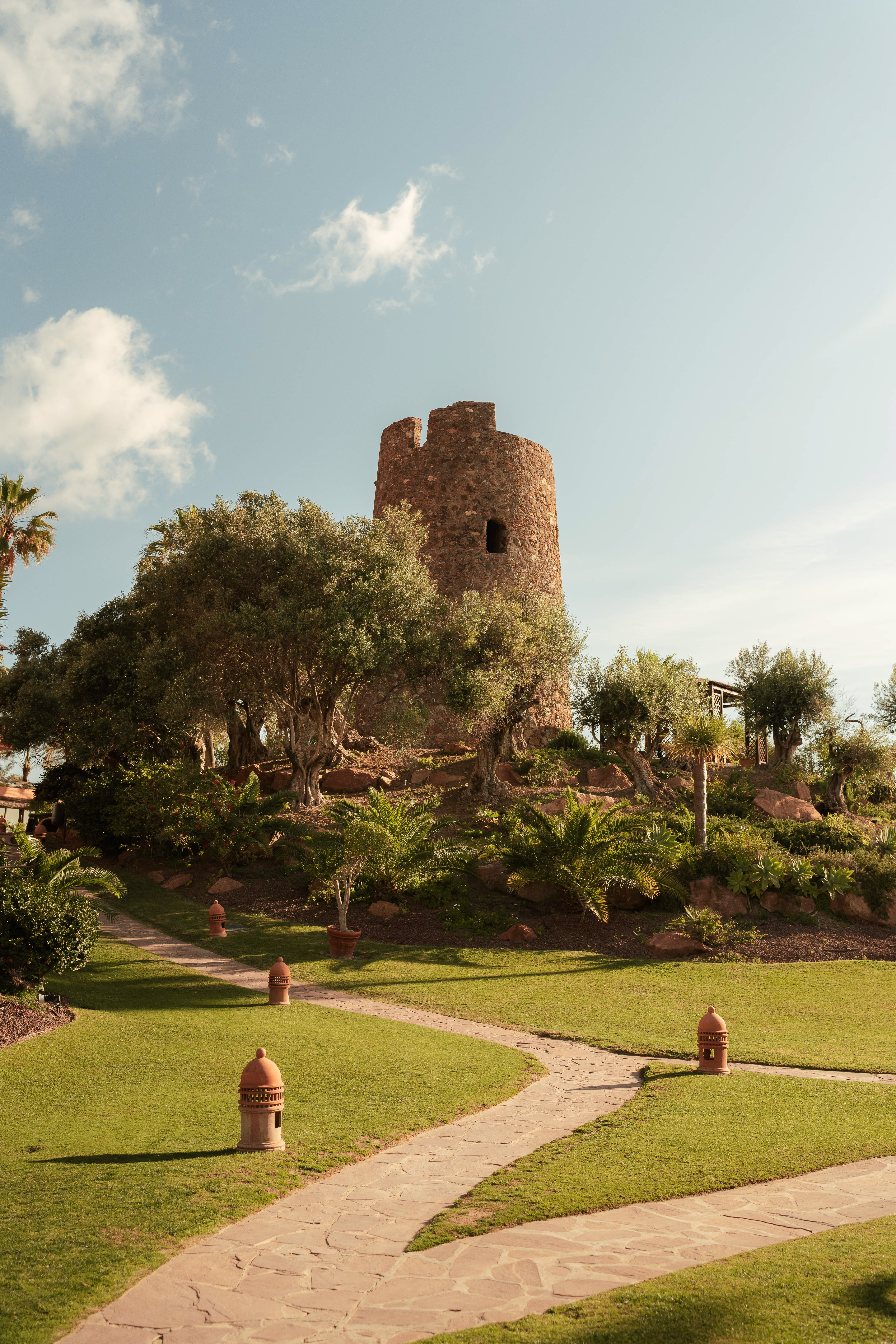 Old stone tower, winding path through a green garden with olive trees.