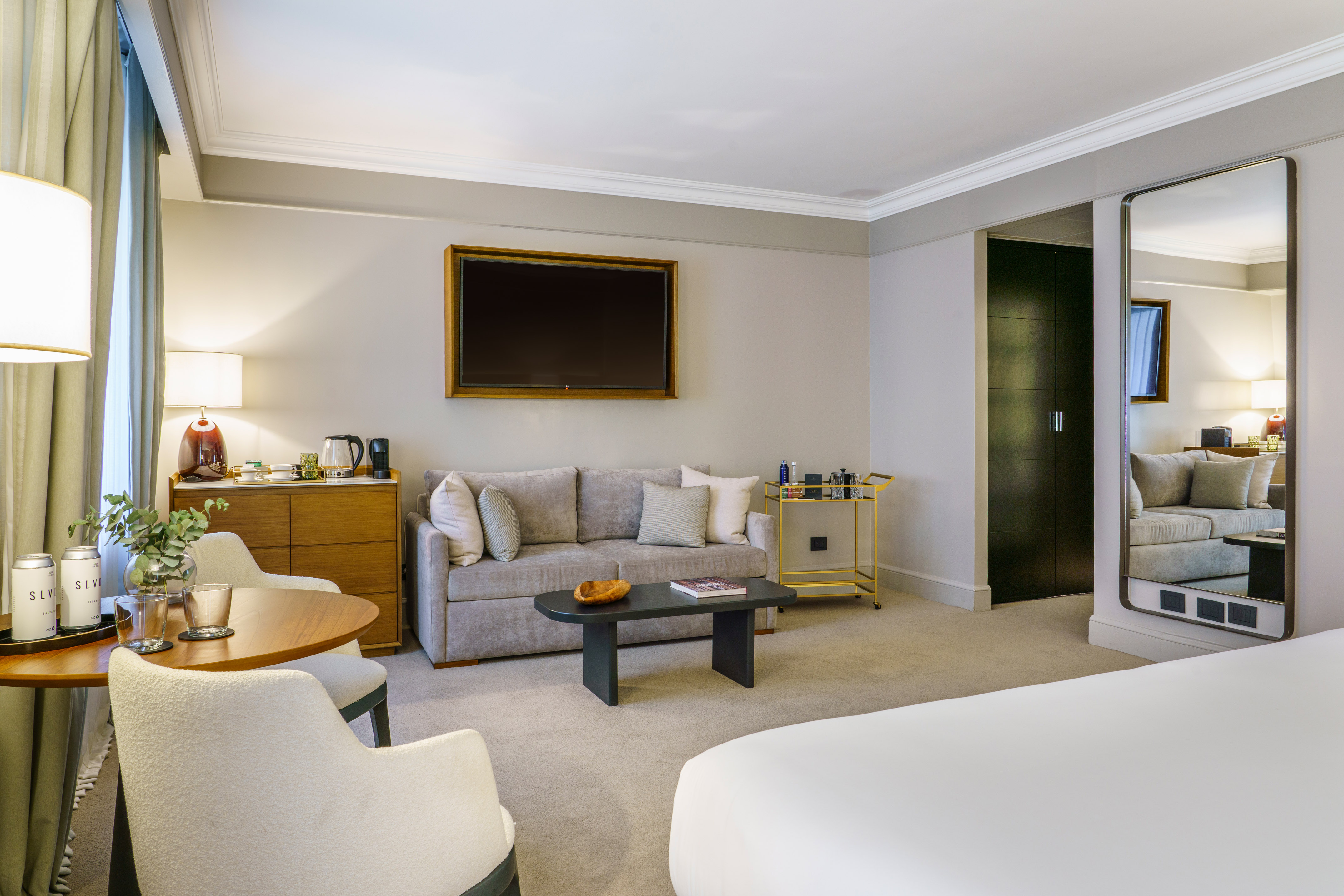 Spacious hotel suite with modern living area, gray sofa, dining table, and large bed.