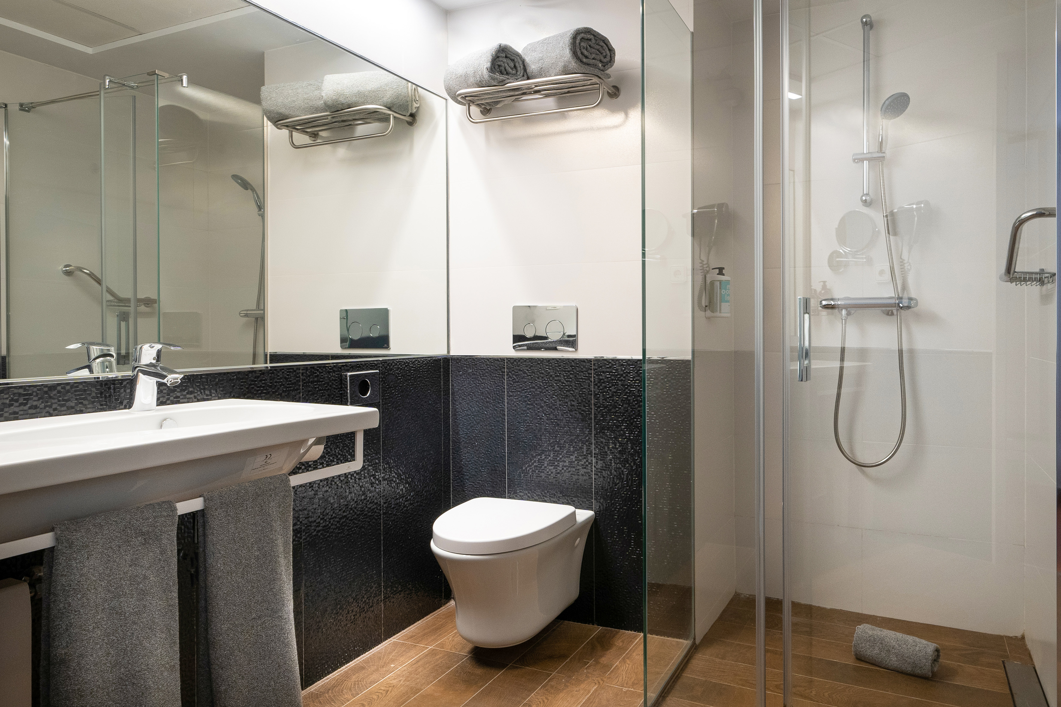 Modern hotel bathroom with glass shower, white sink, toilet, and black tiles.