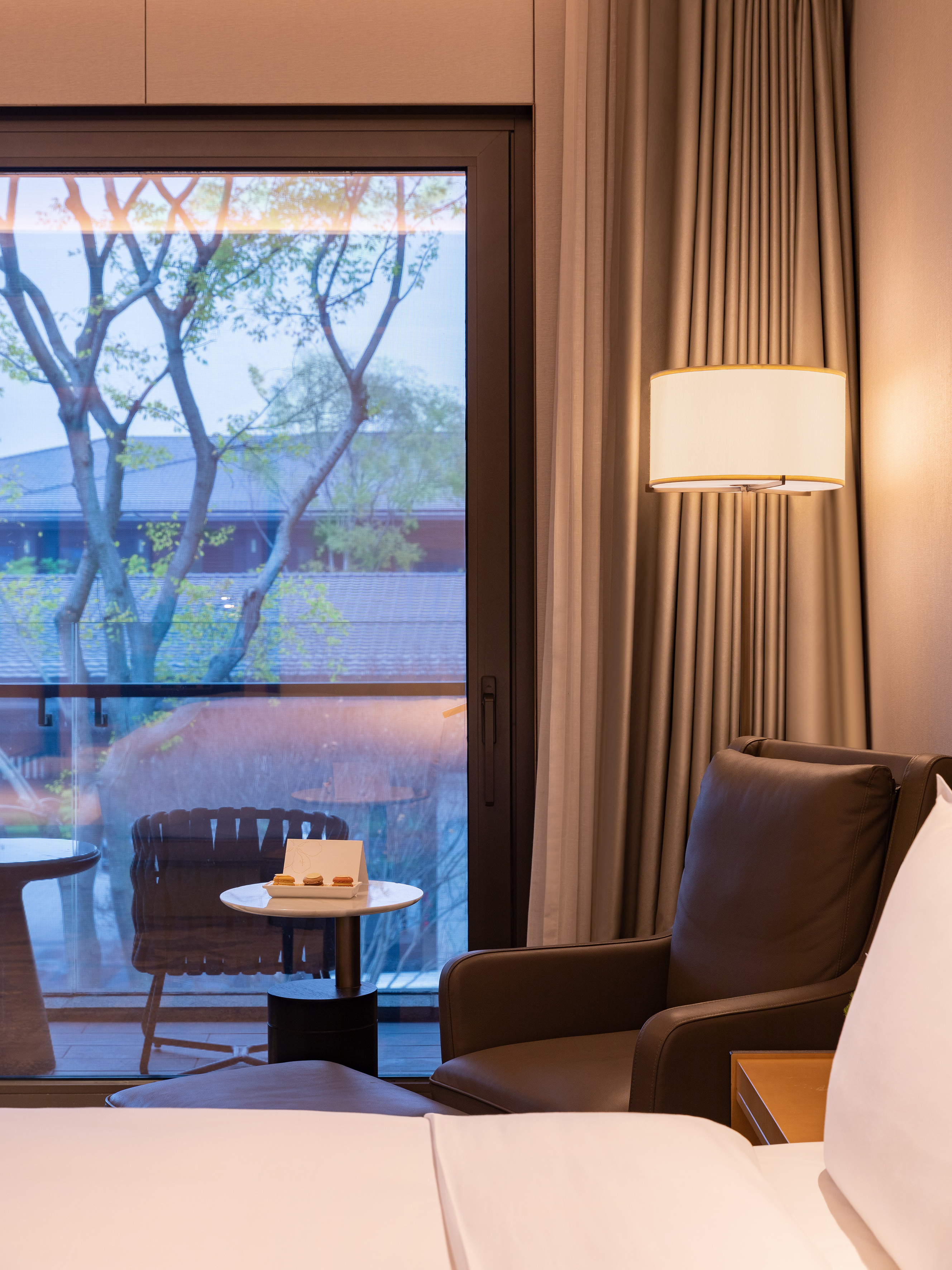 Luxury hotel room with balcony view, leather armchair, floor lamp, and macarons.