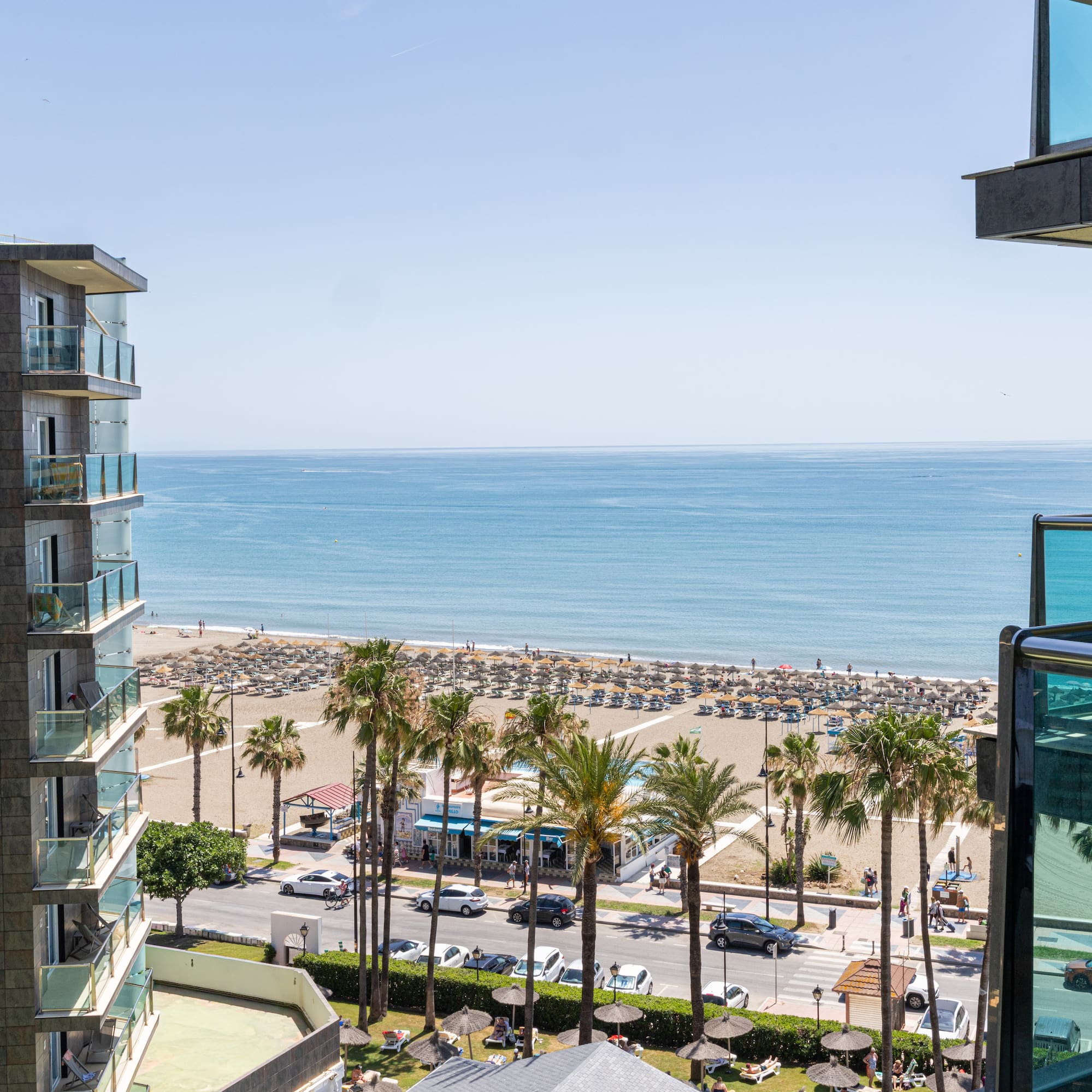 Hotel balcony view of sunny beach, ocean, palm trees, and coastal road.