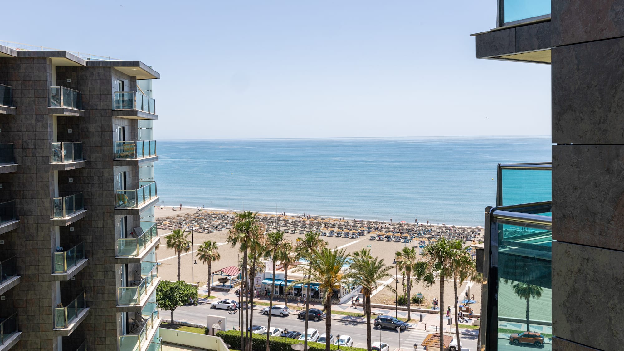Hotel balcony view of sunny beach, ocean, palm trees, and coastal road.