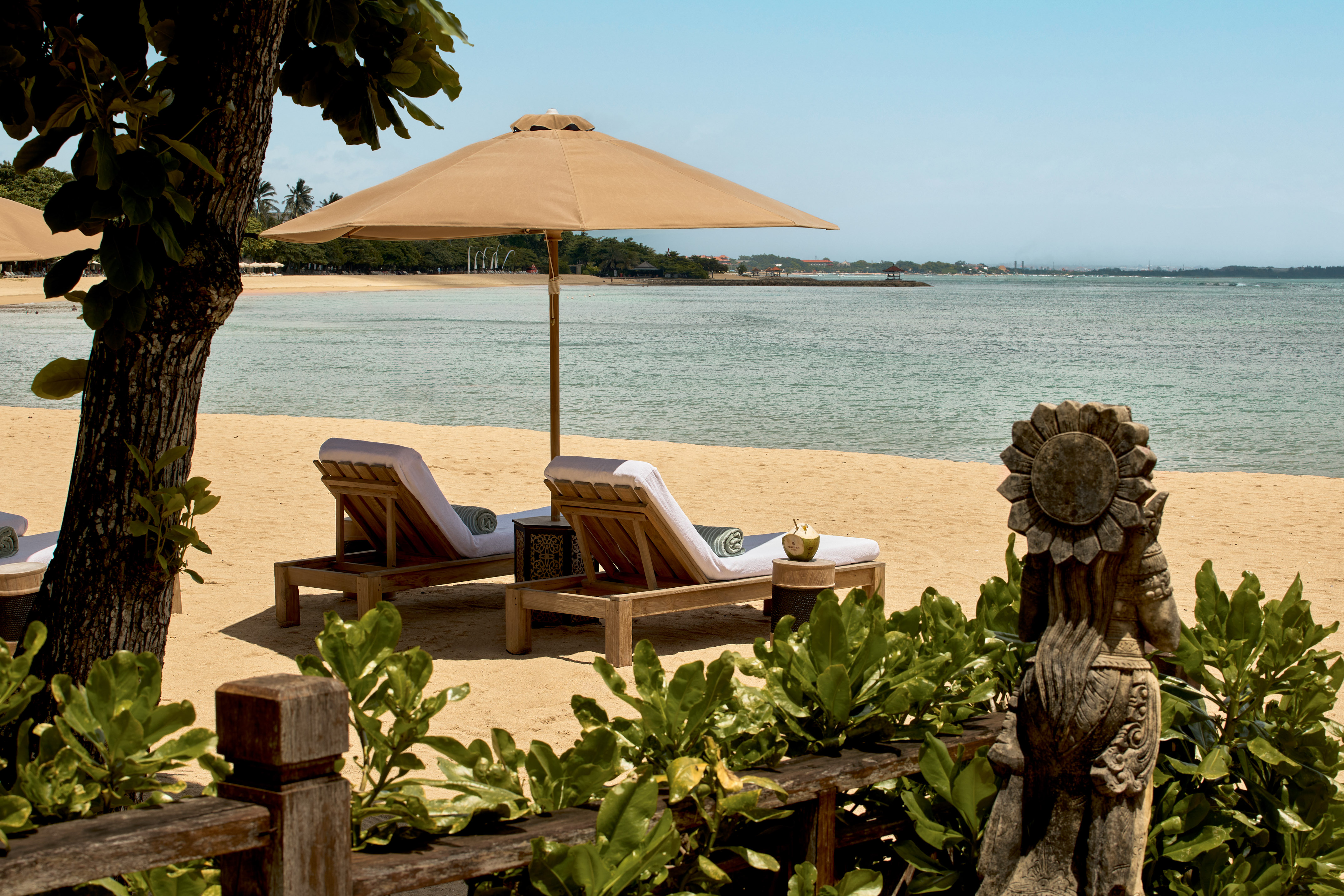 Tropical resort beach with lounge chairs, umbrella, ocean, and Balinese statue.