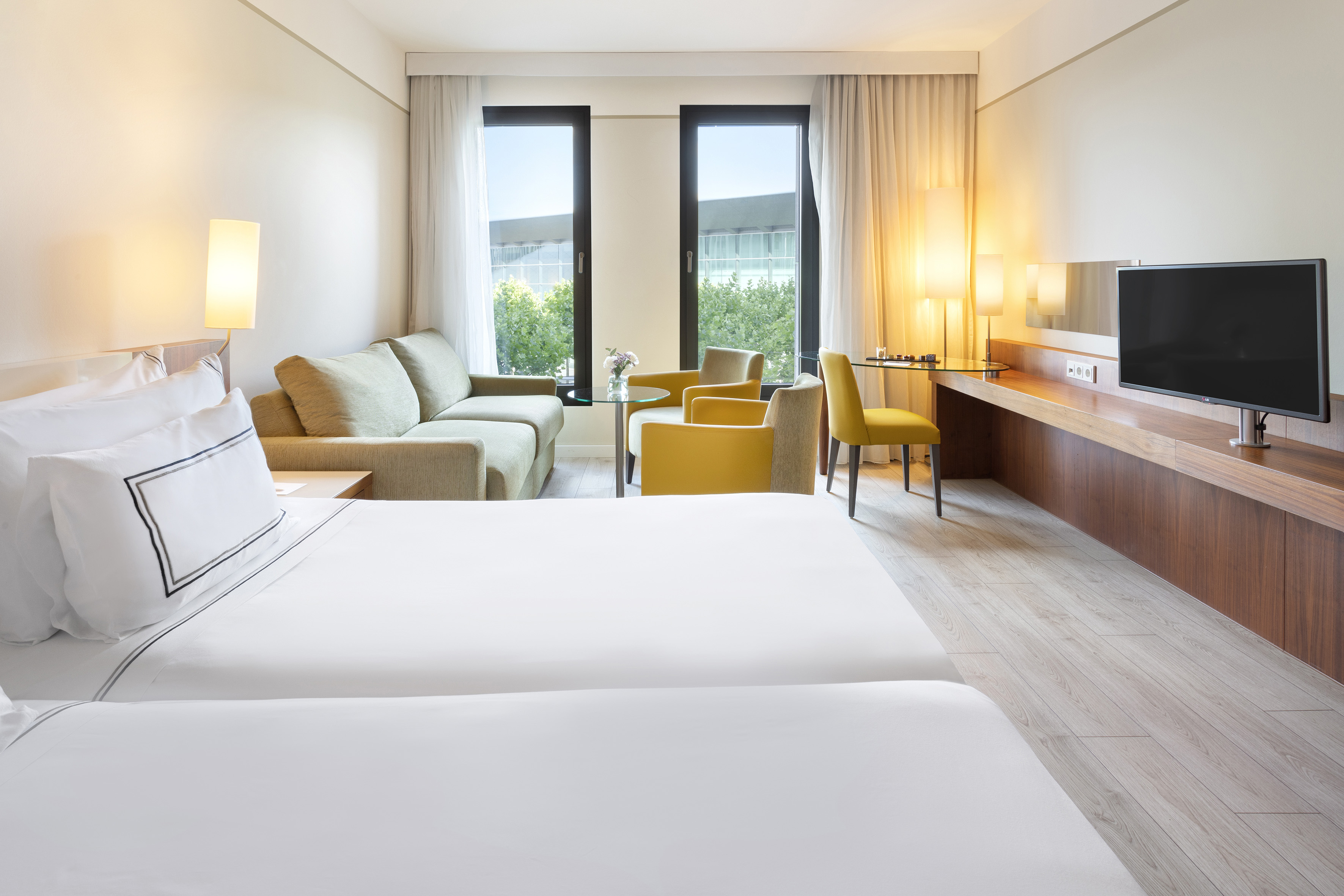 Modern hotel guest room with two beds, sofa, desk, TV, and bright windows.