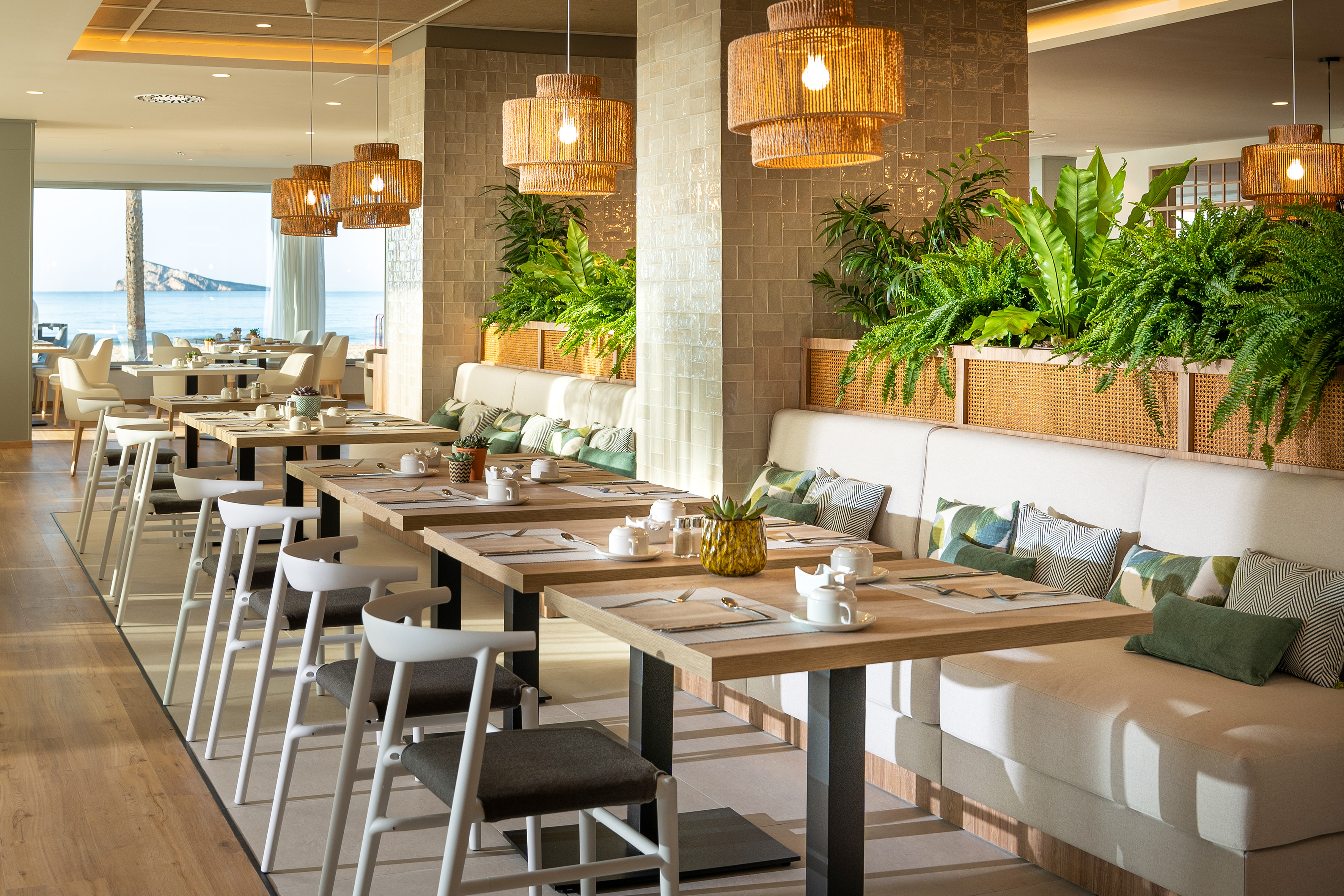 Modern restaurant with ocean view, rattan lights, green plants, and set tables.