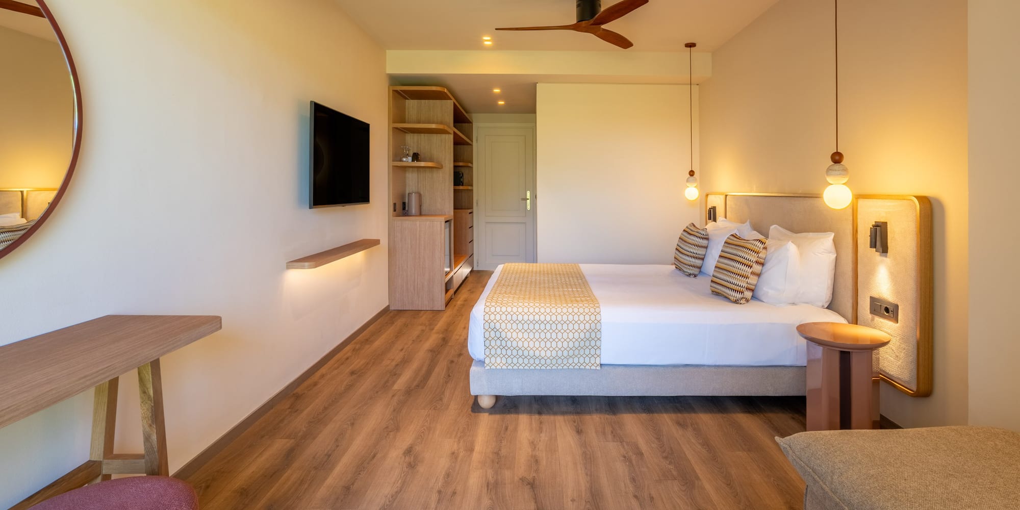 Contemporary hotel bedroom with large bed, TV, wooden floor, ceiling fan.