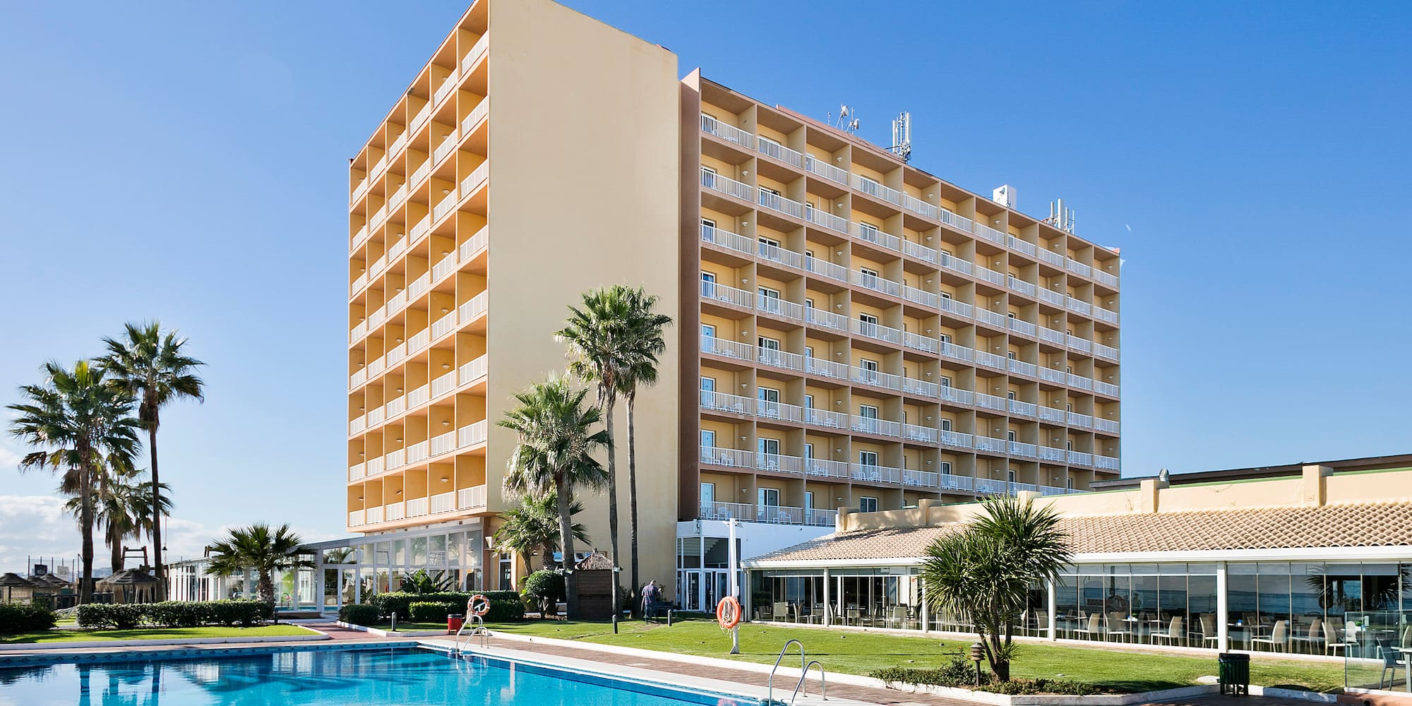 Sunny resort hotel with large swimming pool, palm trees, and many balconies.