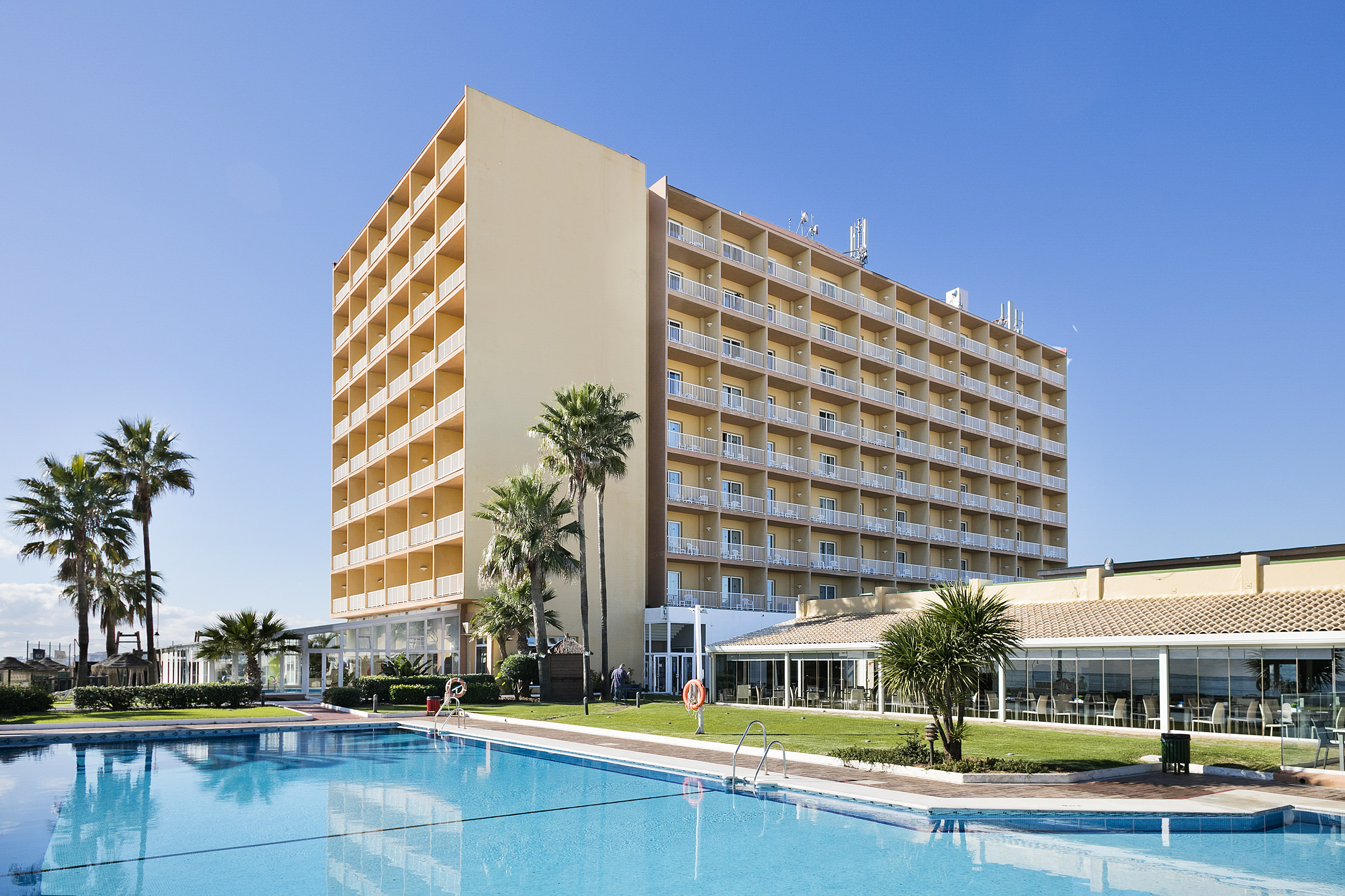 Sunny resort hotel with large swimming pool, palm trees, and many balconies.