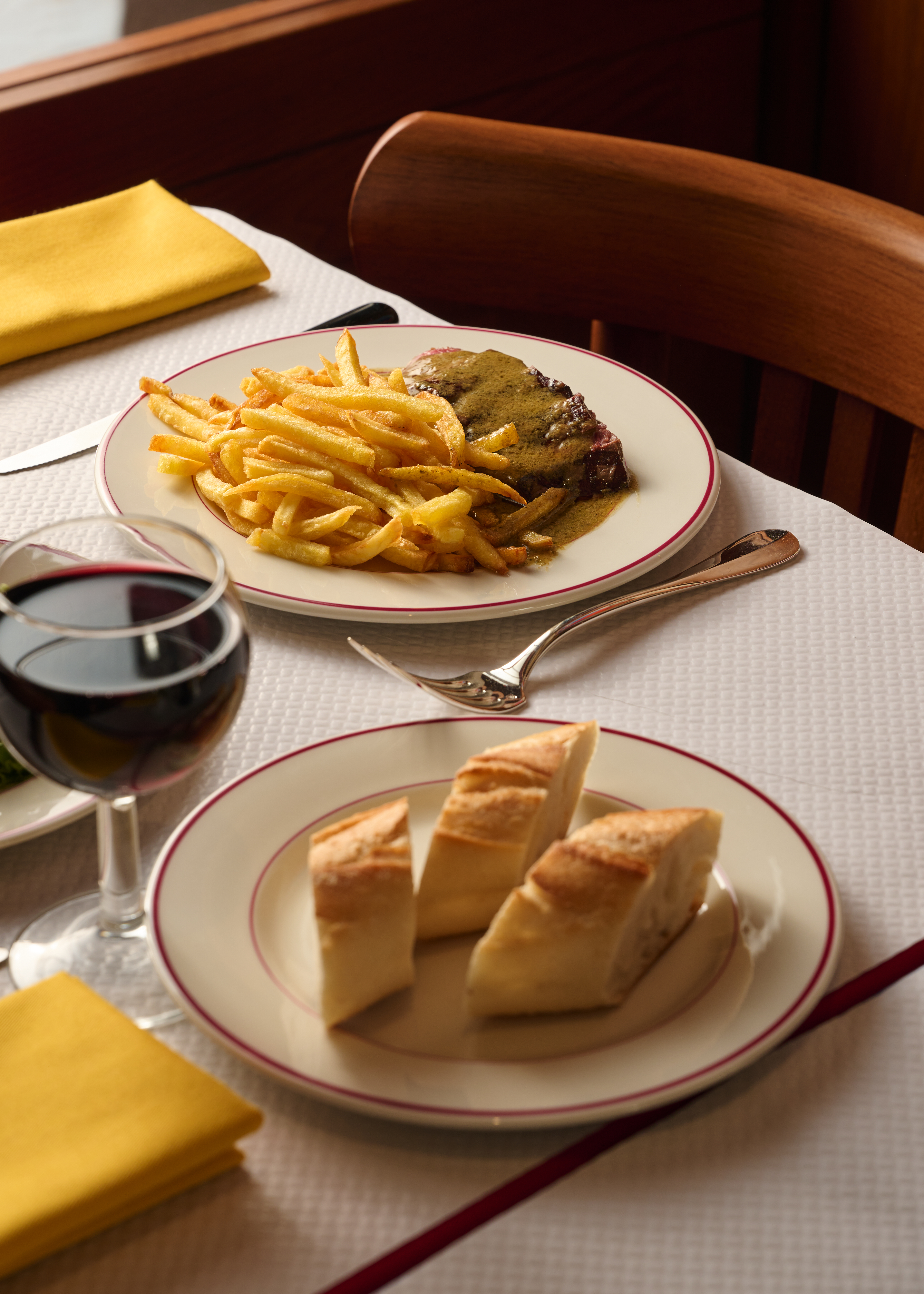 Steak frites with green sauce, baguette, and red wine in a restaurant setting.