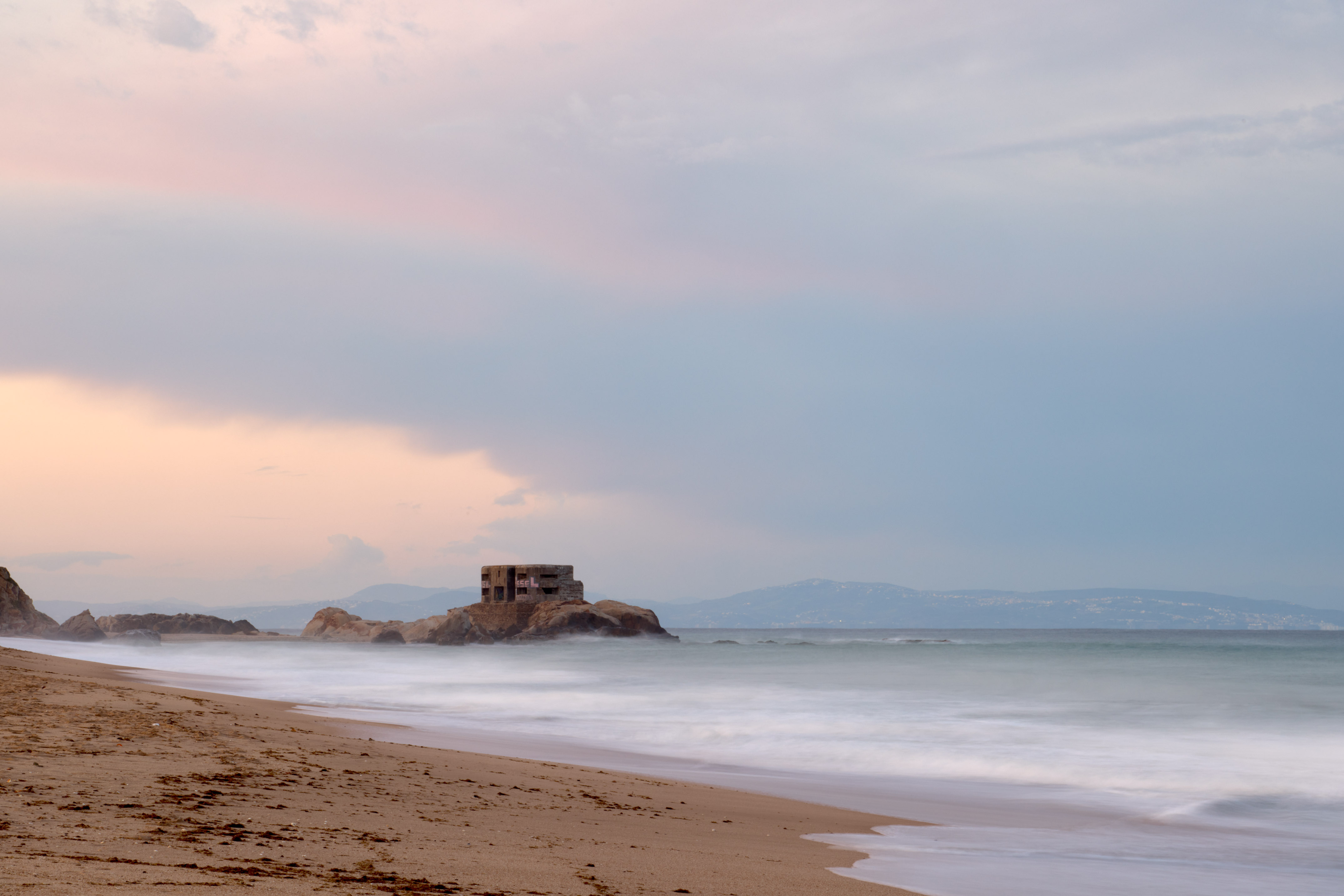 Coastal bunker on a rocky island, long exposure ocean, sandy beach at sunset.