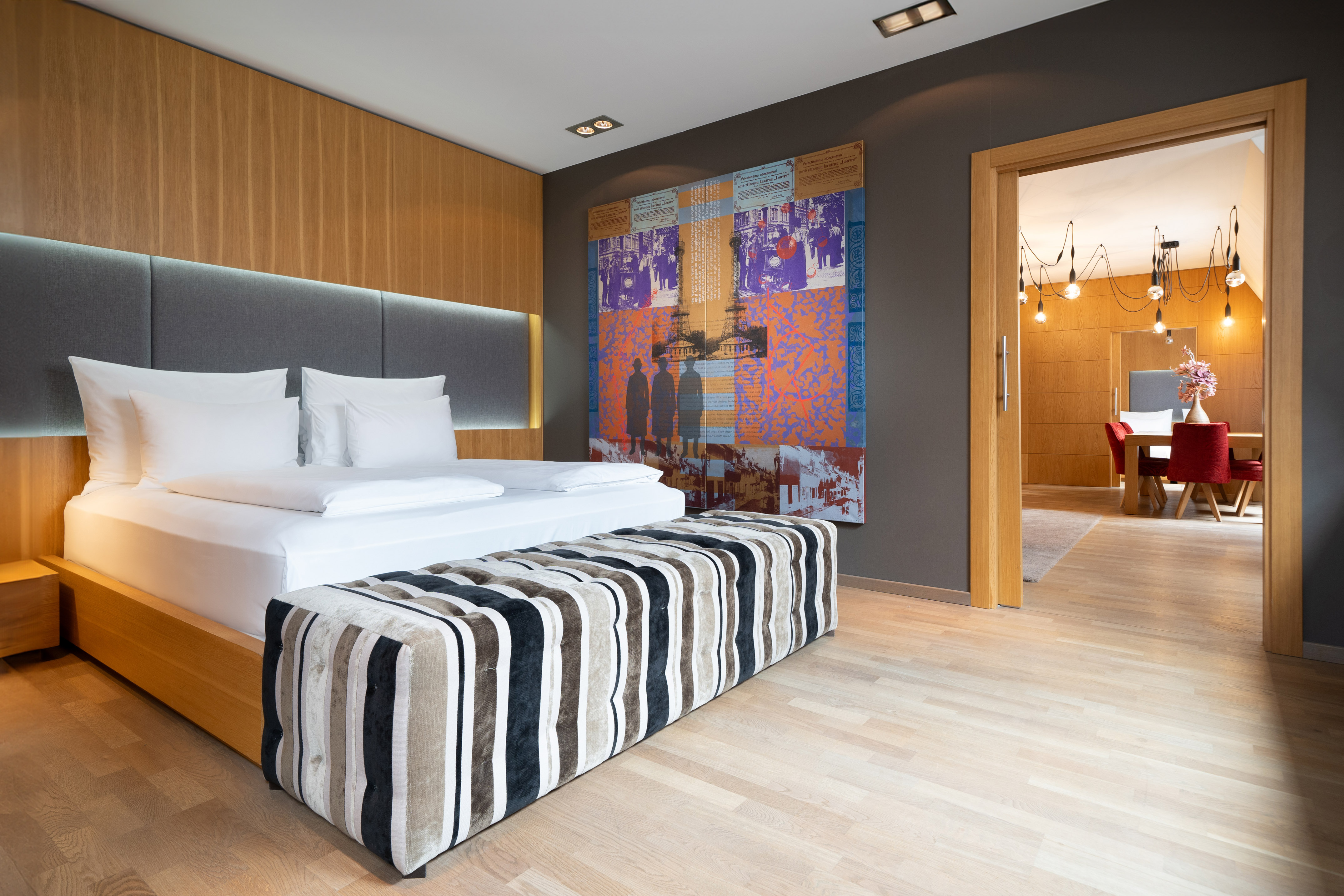 Modern hotel room with bed, striped ottoman, abstract art, and wood interior.