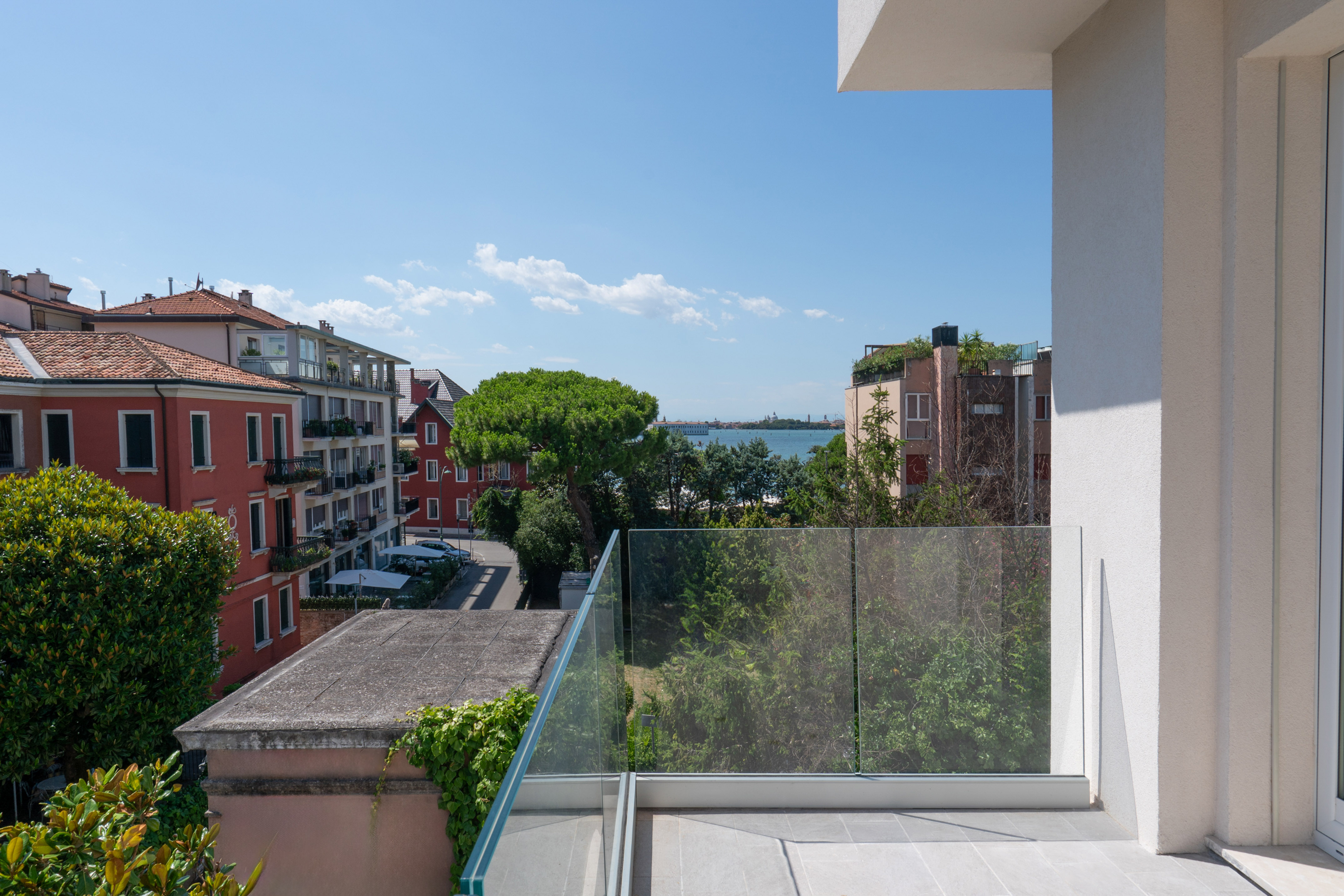 Panoramic balcony view of city buildings, green trees, and distant sea.