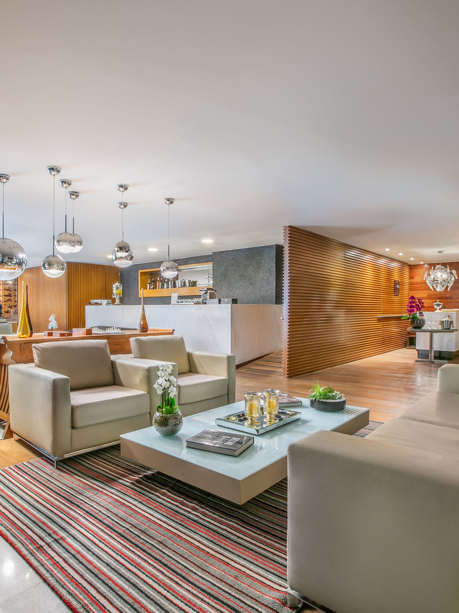 Modern luxury lounge with beige sofas, glass table, wood wall, and integrated bar.