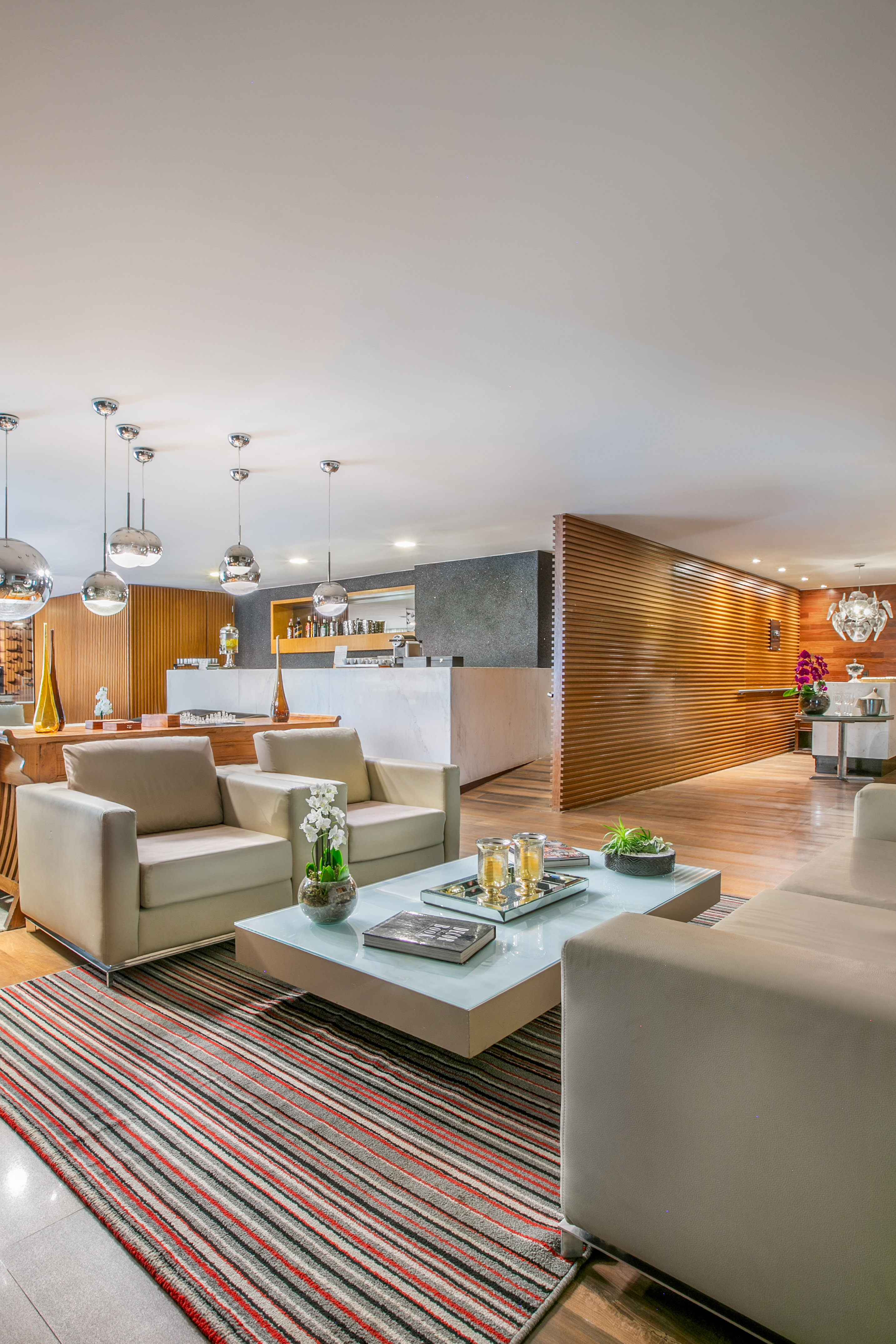 Modern luxury lounge with beige sofas, glass table, wood wall, and integrated bar.