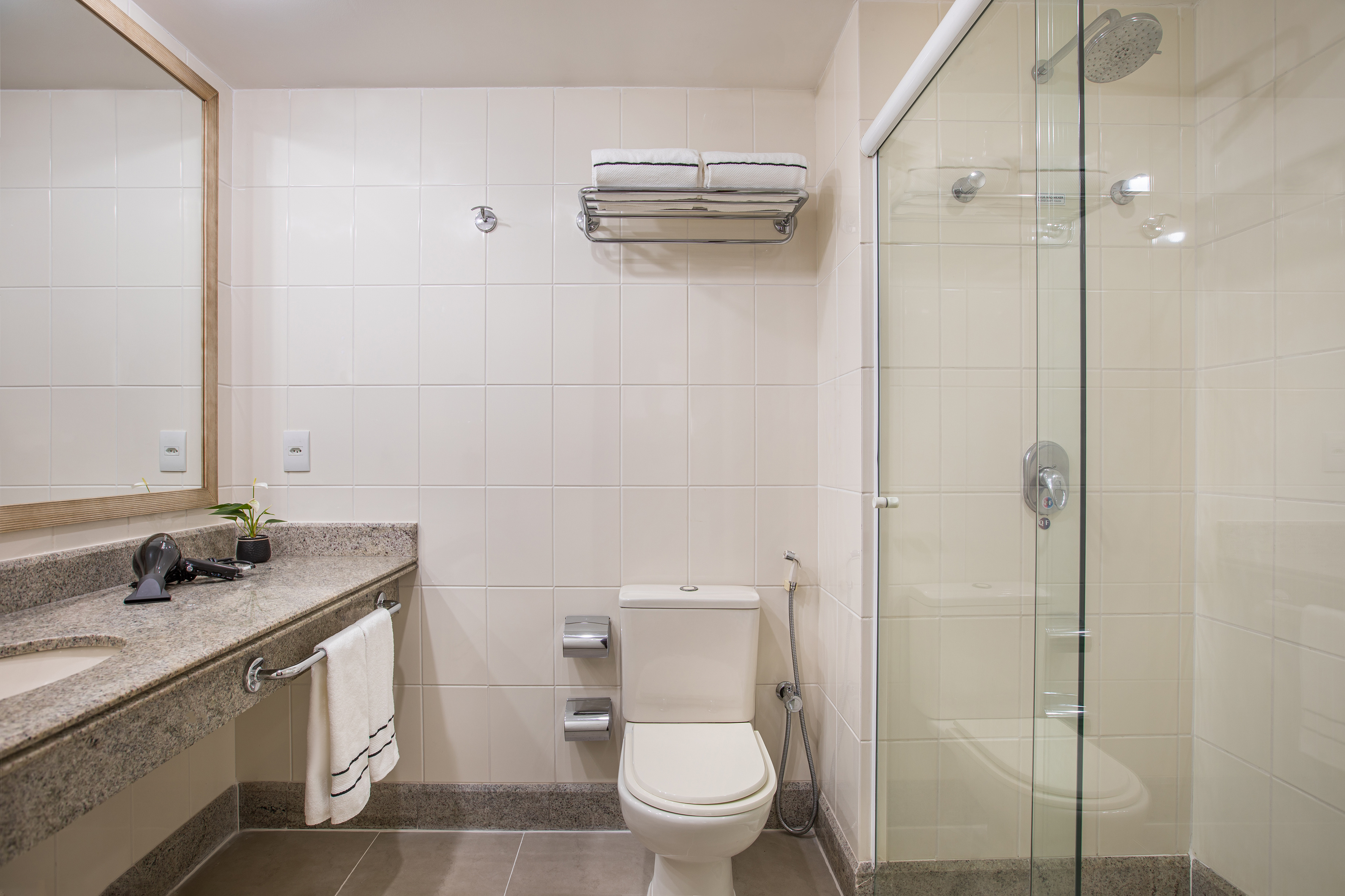 Hotel bathroom with white tiles, granite vanity, toilet, and glass shower.