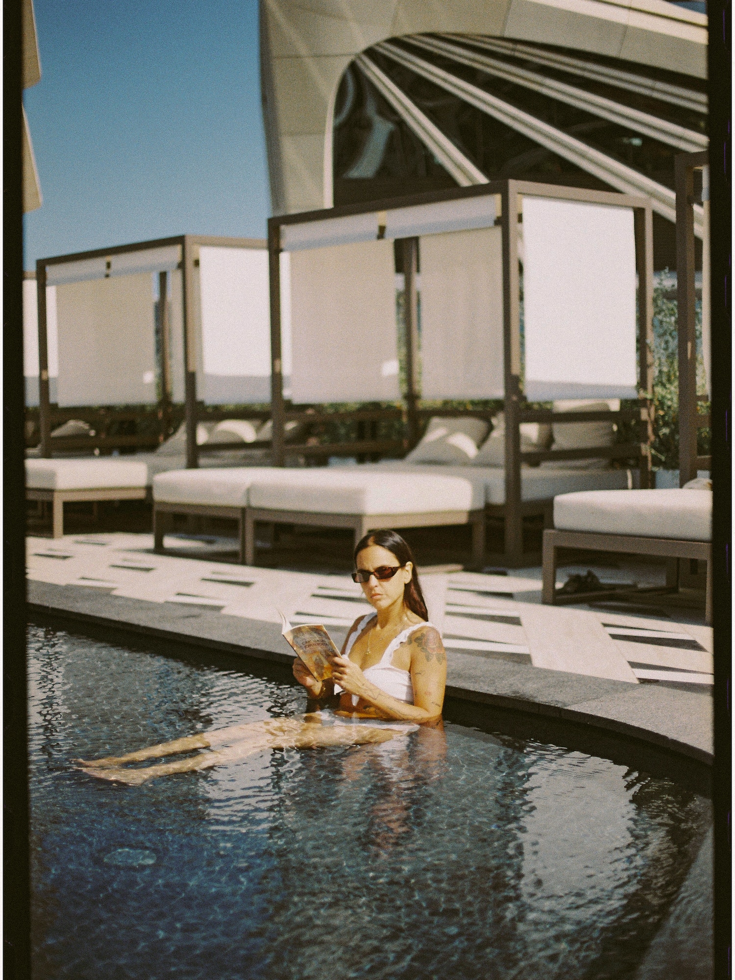 Woman in sunglasses reading a book in a luxury outdoor pool with cabanas.