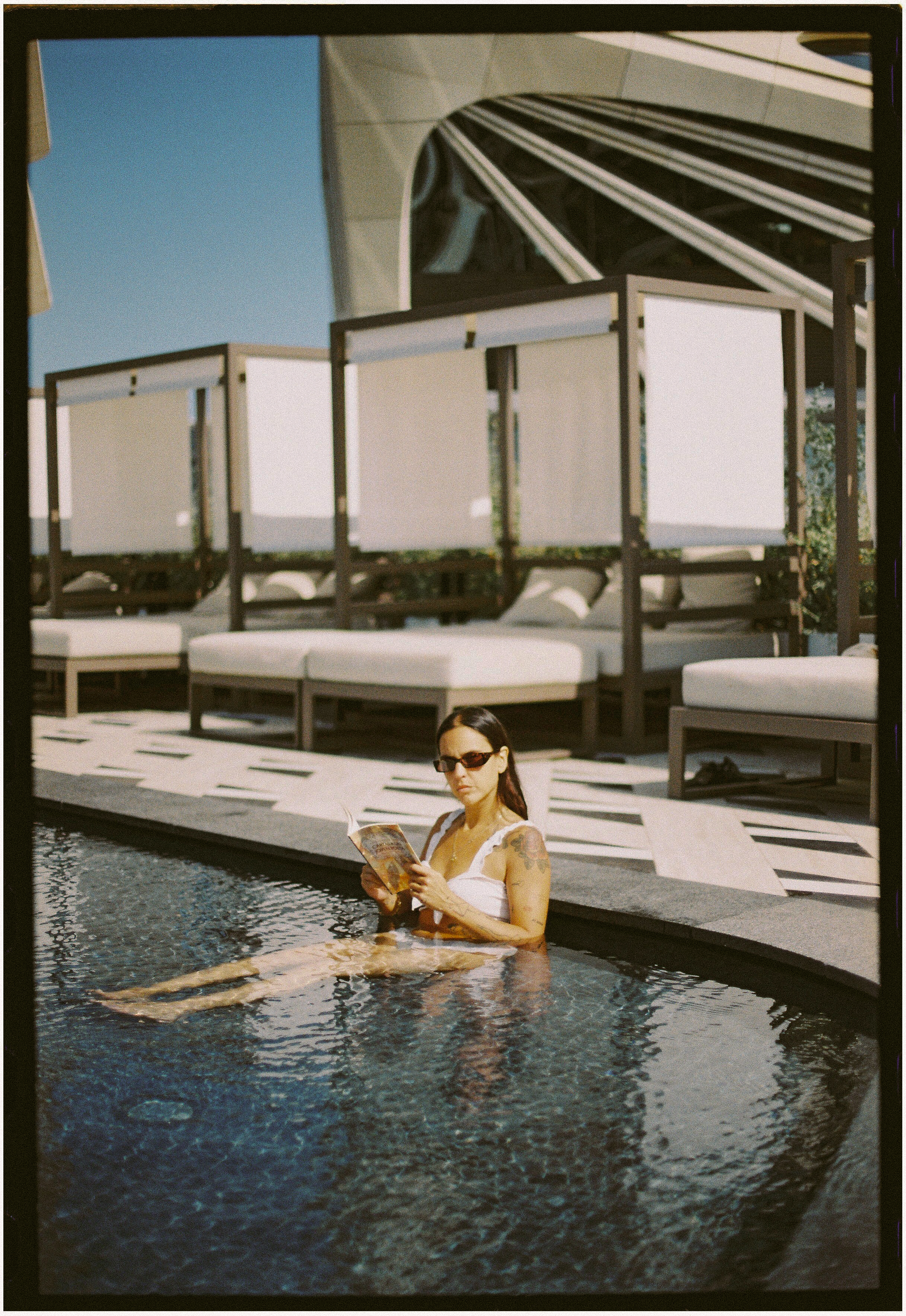 Woman in sunglasses reading a book in a luxury outdoor pool with cabanas.