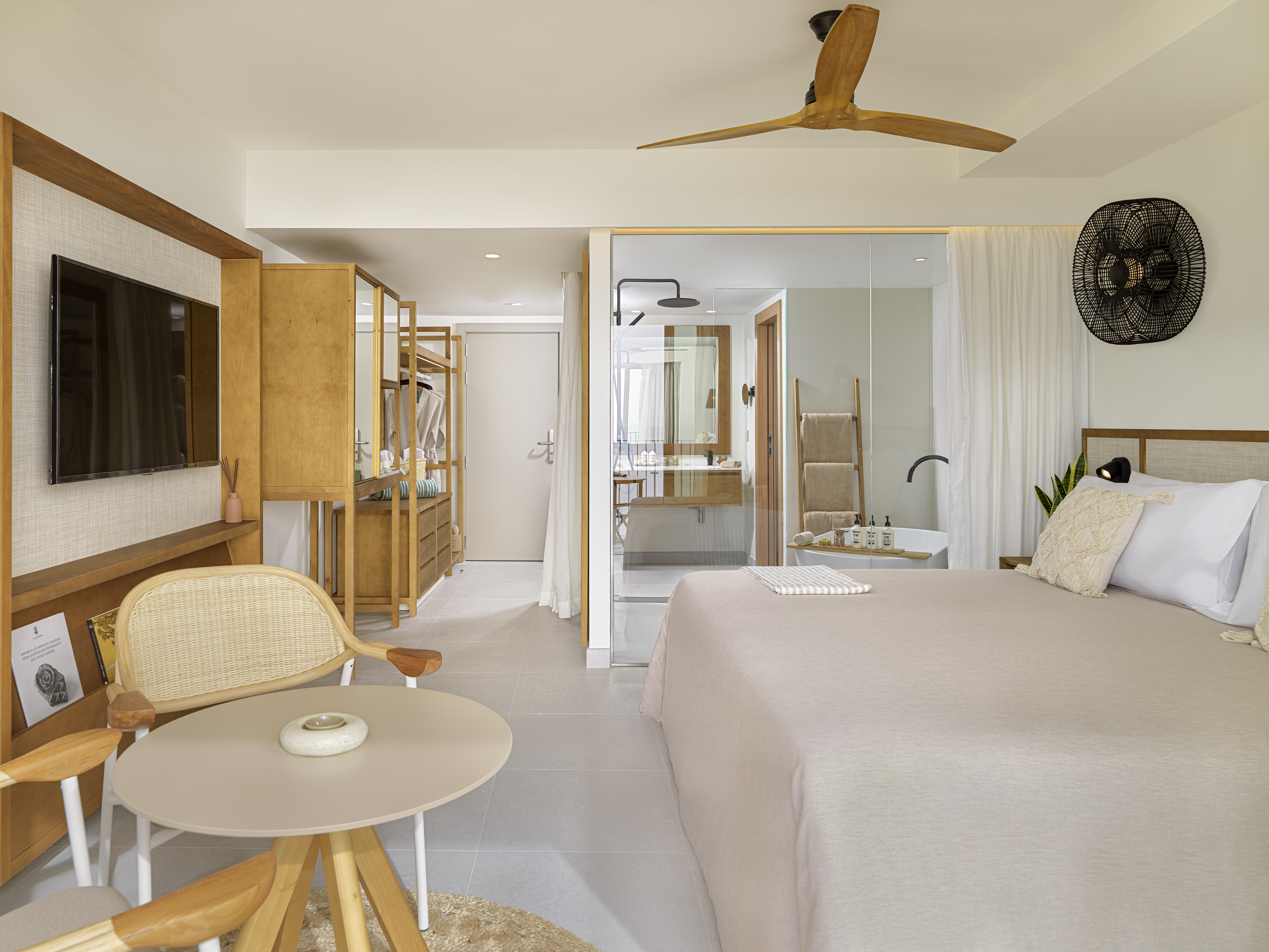 Modern hotel room: king bed, open concept bathroom, wooden fan, rattan chair.