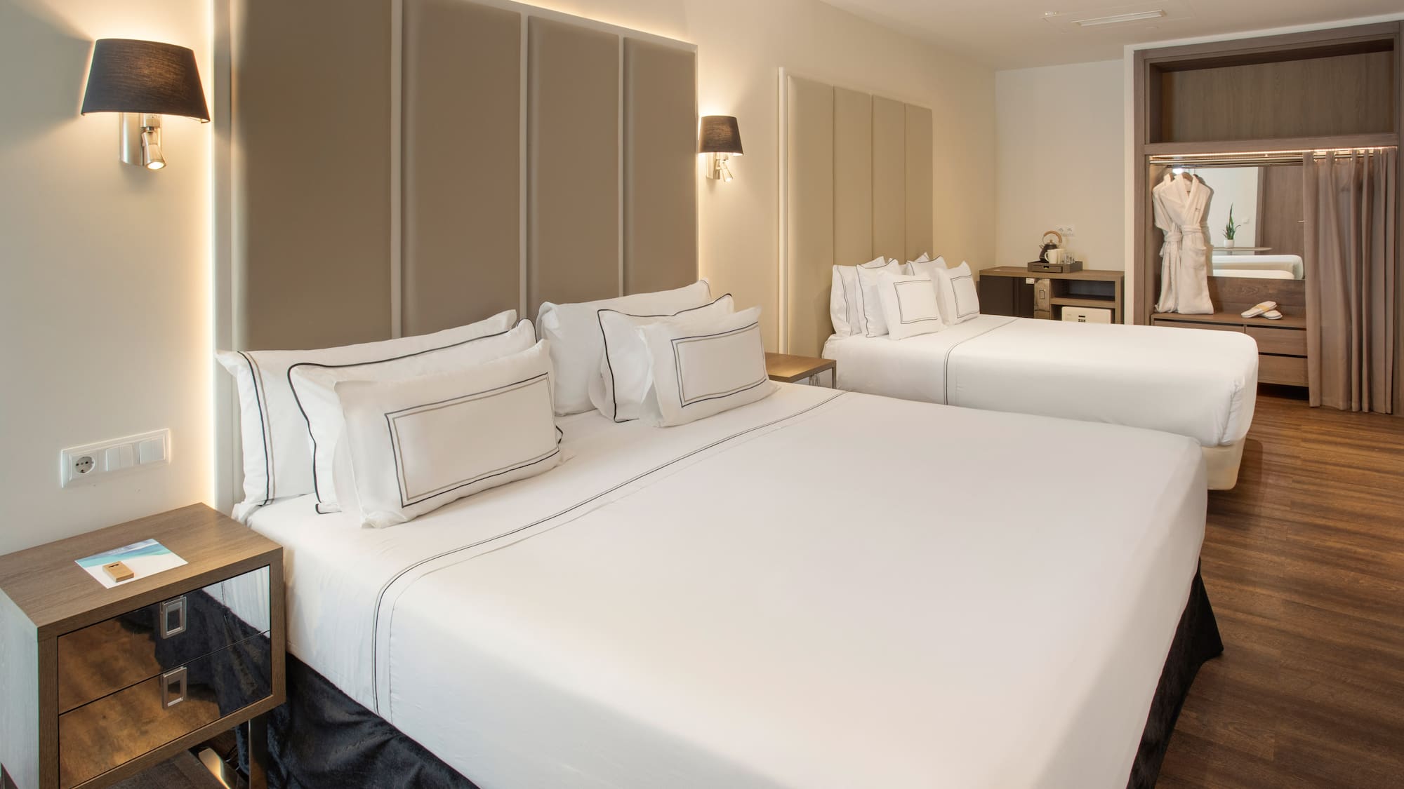Modern hotel room with two beds, white bedding, upholstered headboards, and a wardrobe.