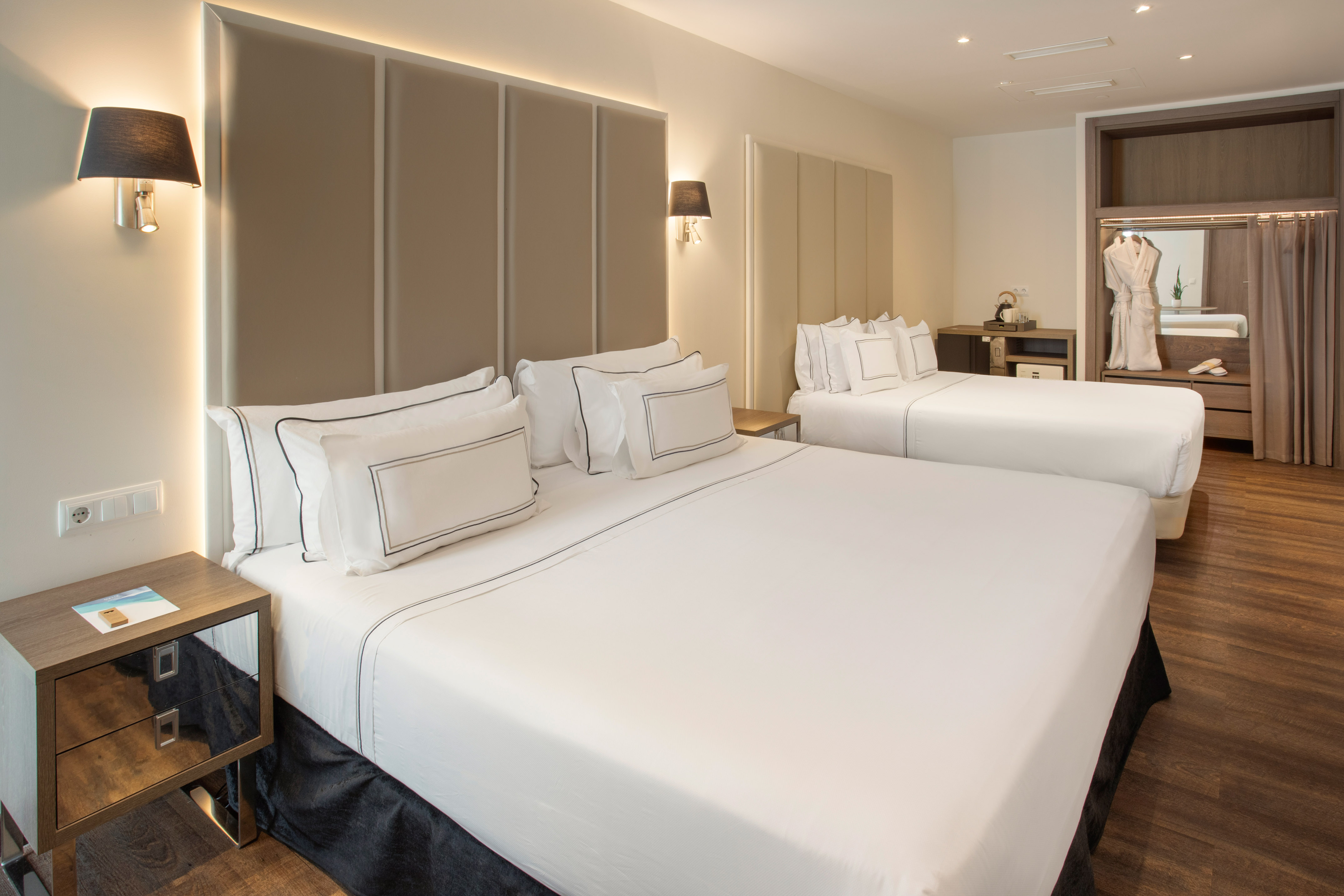 Modern hotel room with two beds, white bedding, upholstered headboards, and a wardrobe.