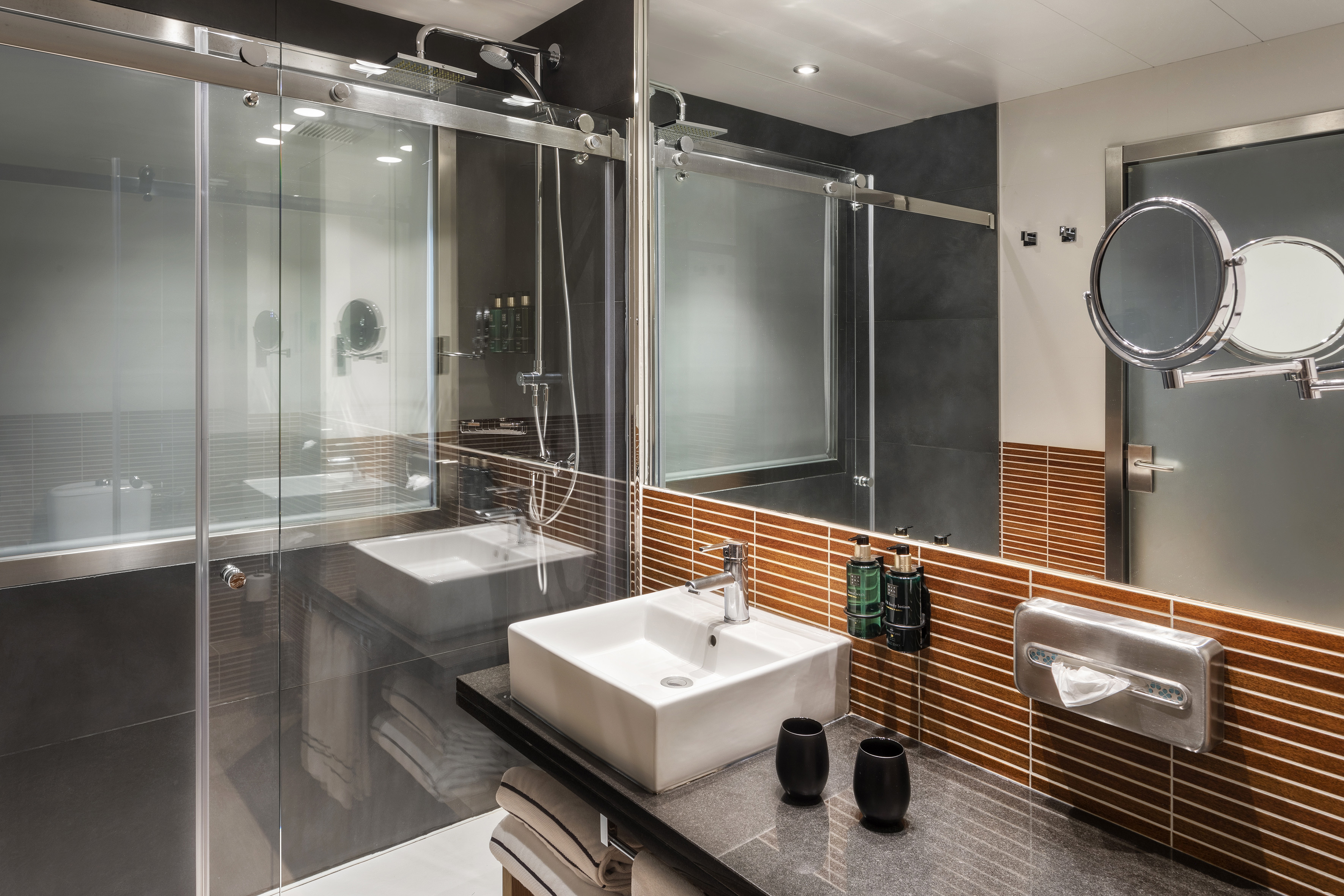 Modern hotel bathroom with glass shower, white sink, and brown tiled wall.
