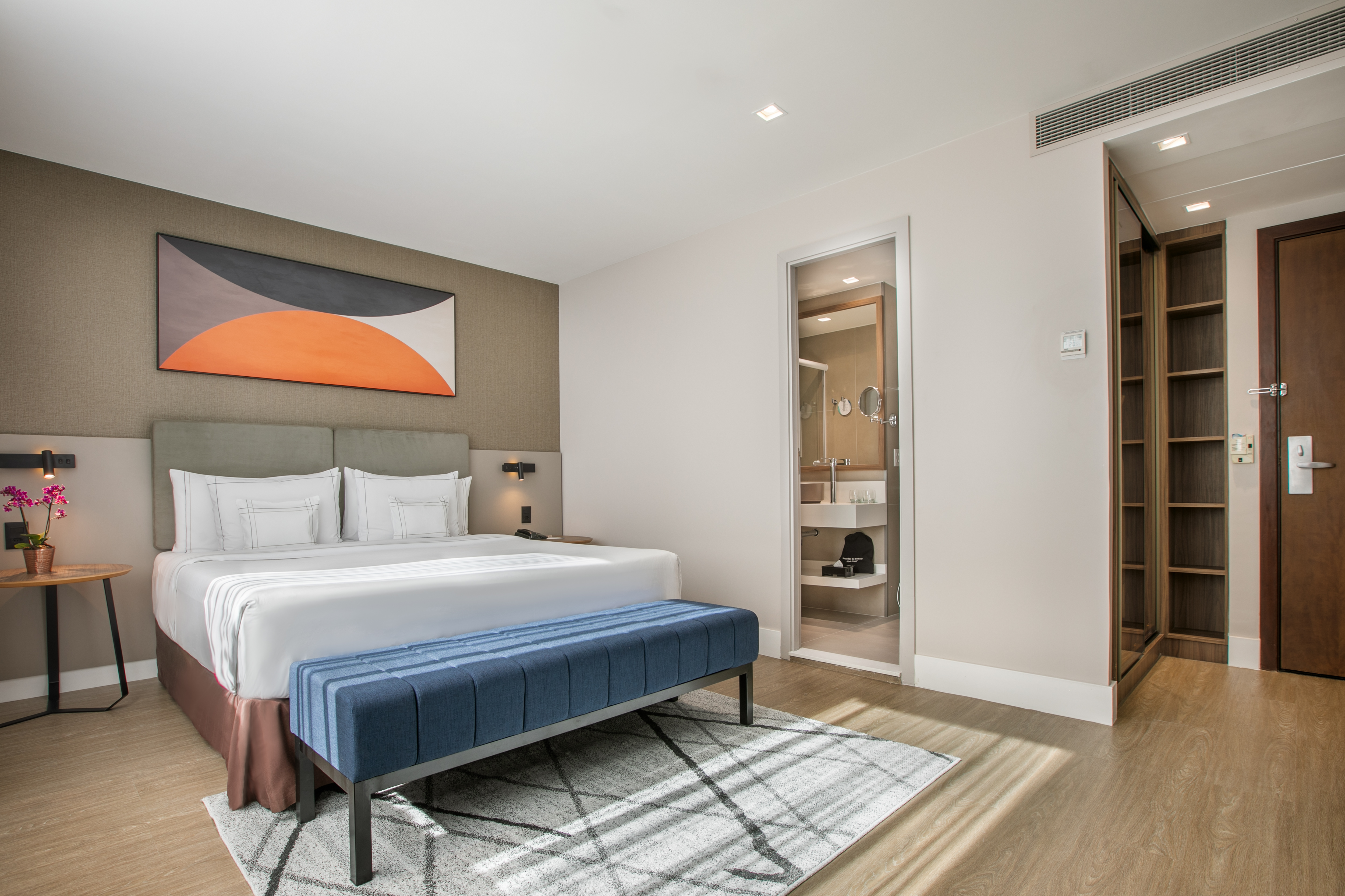 Modern hotel bedroom with large bed, blue bench, abstract art, and bathroom.