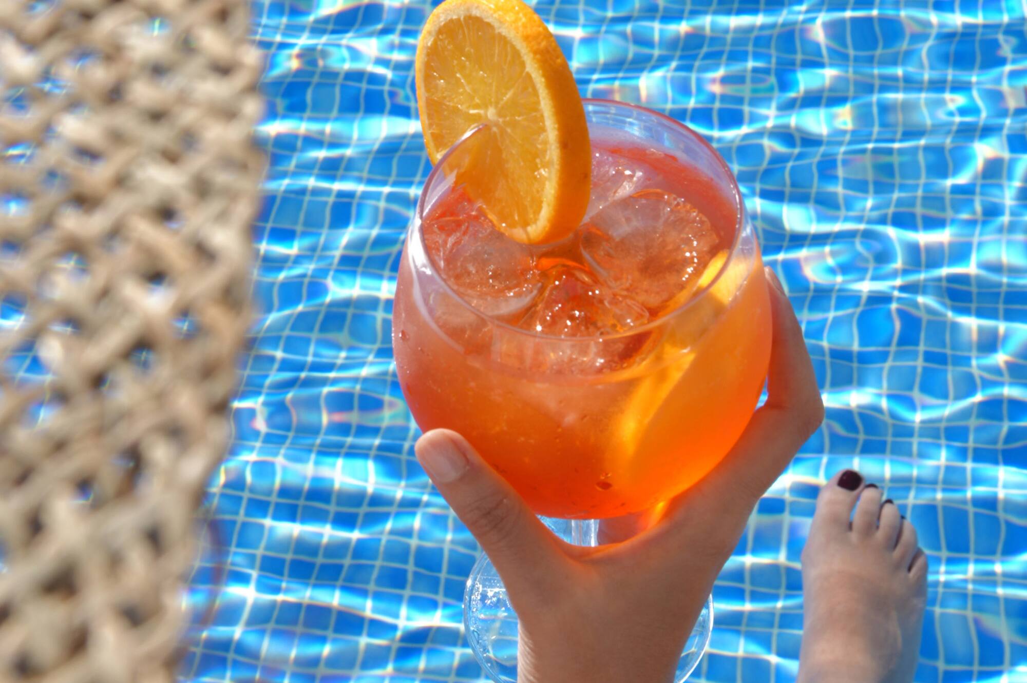 Hand holding orange cocktail with orange slice, legs submerged in blue pool.