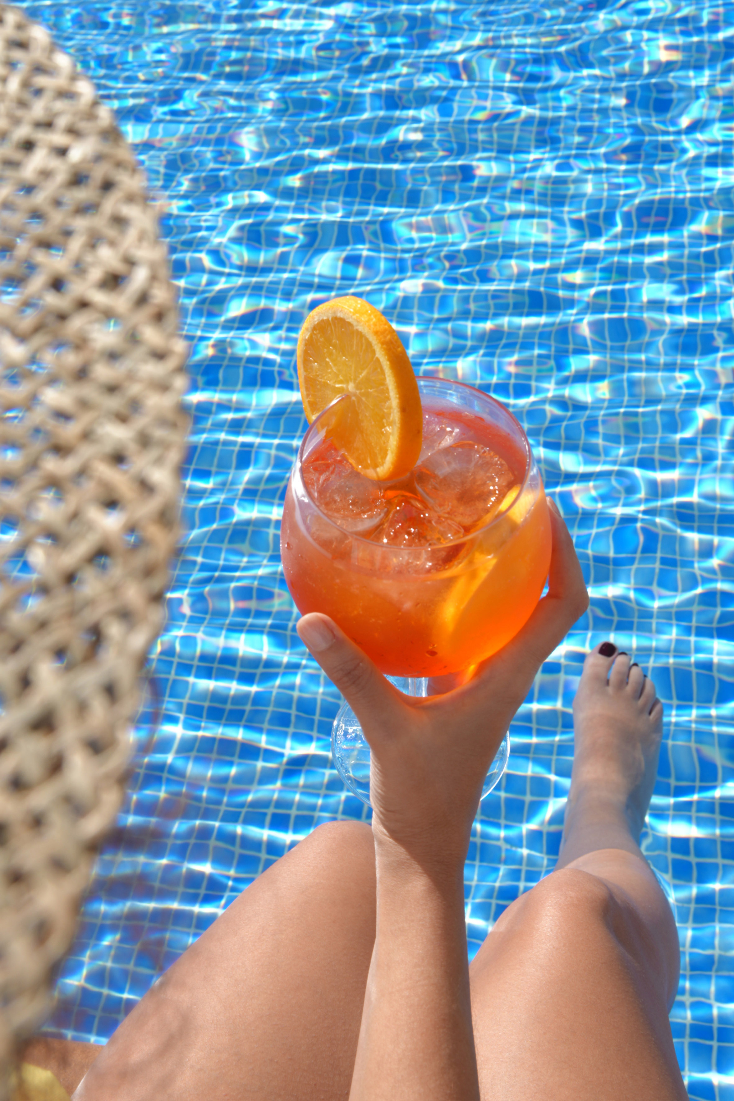 Hand holding orange cocktail with orange slice, legs submerged in blue pool.