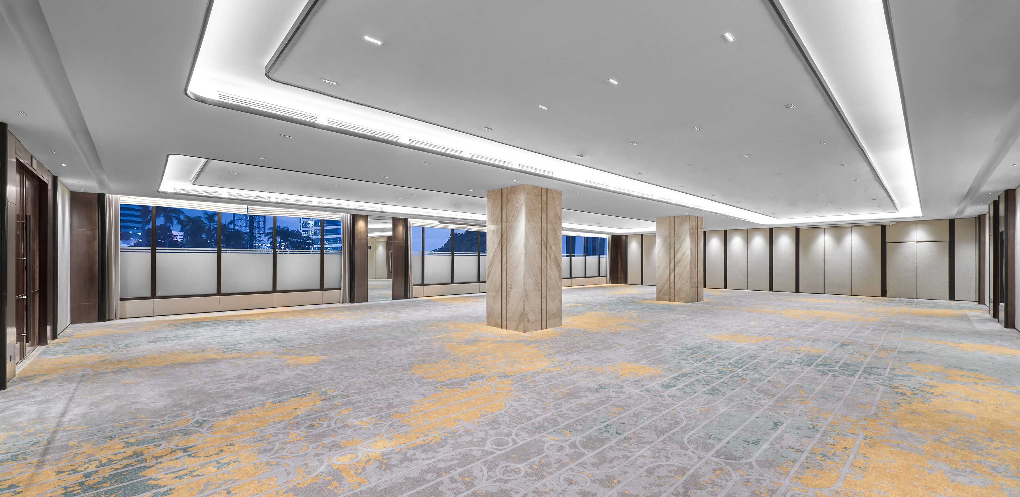 Modern empty ballroom with patterned carpet, marble pillars, and city views.