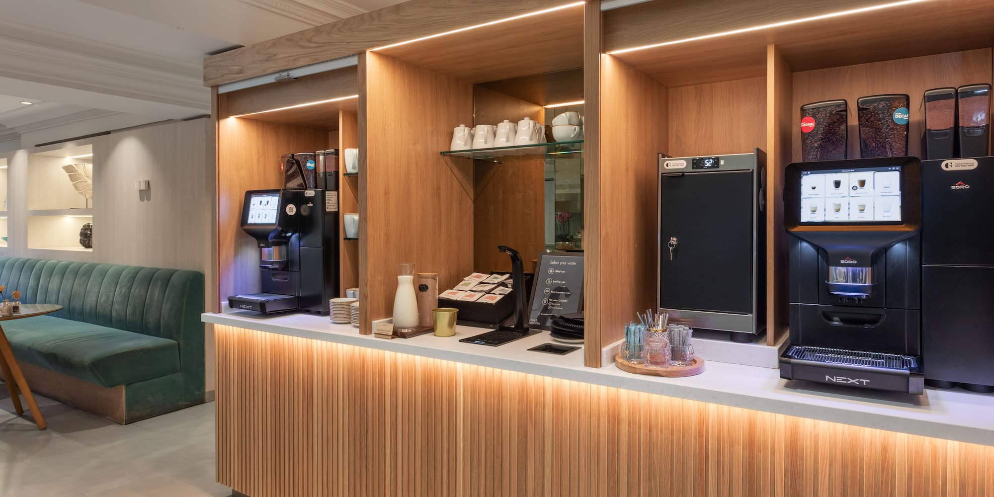 Modern self-service coffee bar with espresso machines, wooden counter, and lounge.