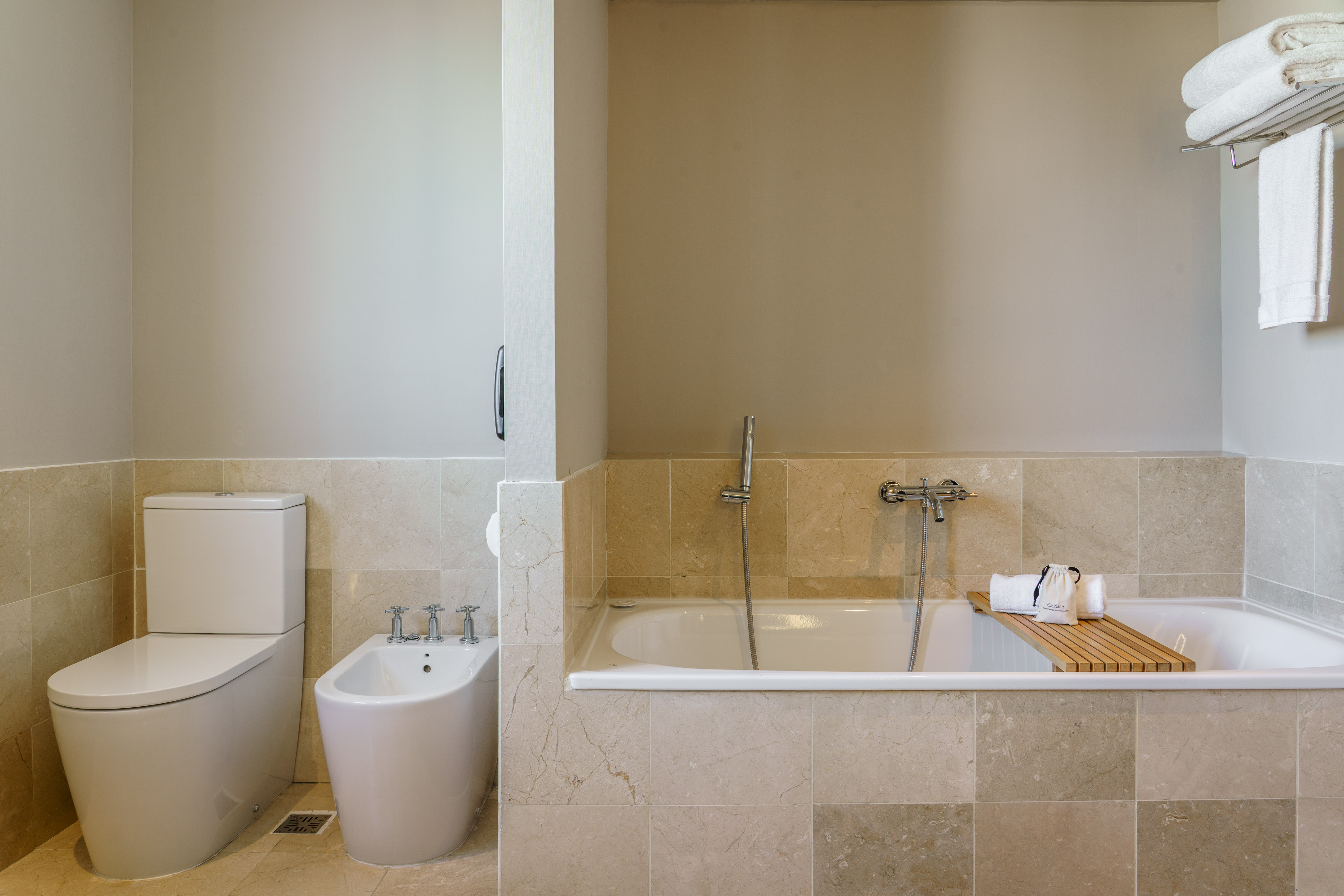 Elegant hotel bathroom with toilet, bidet, bathtub, and beige marble tiles.