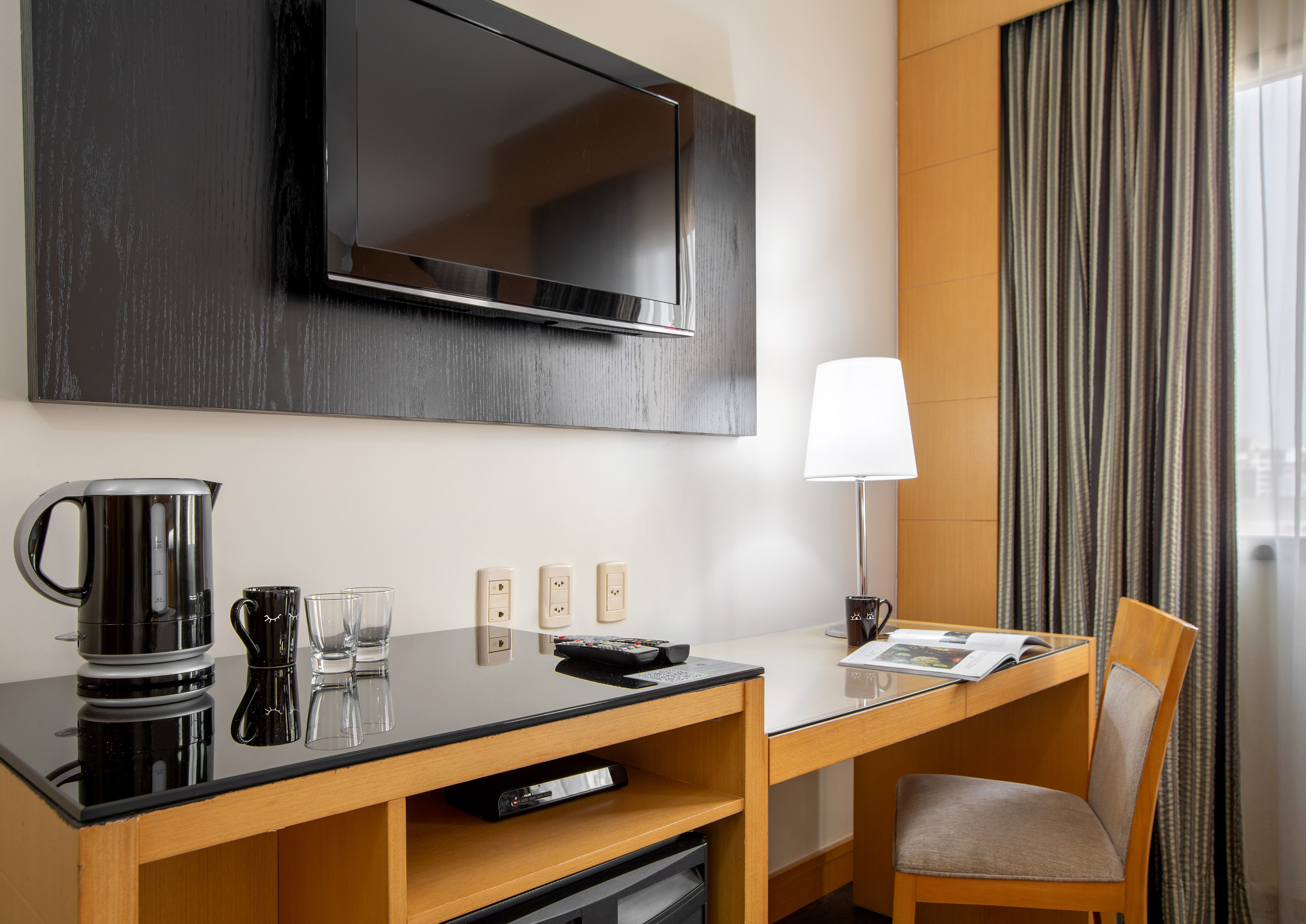 Hotel guest room with wall-mounted TV, wooden desk, chair, kettle, and minibar.