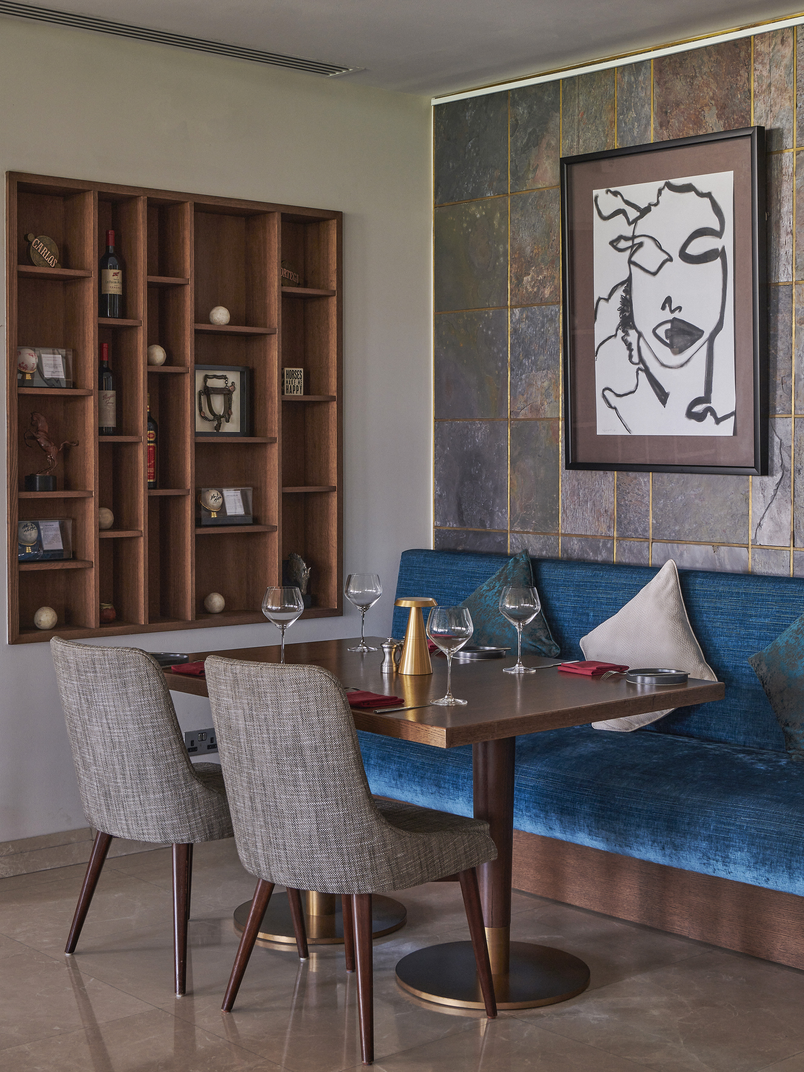 Modern restaurant dining with blue velvet banquette, wooden shelves, abstract art.