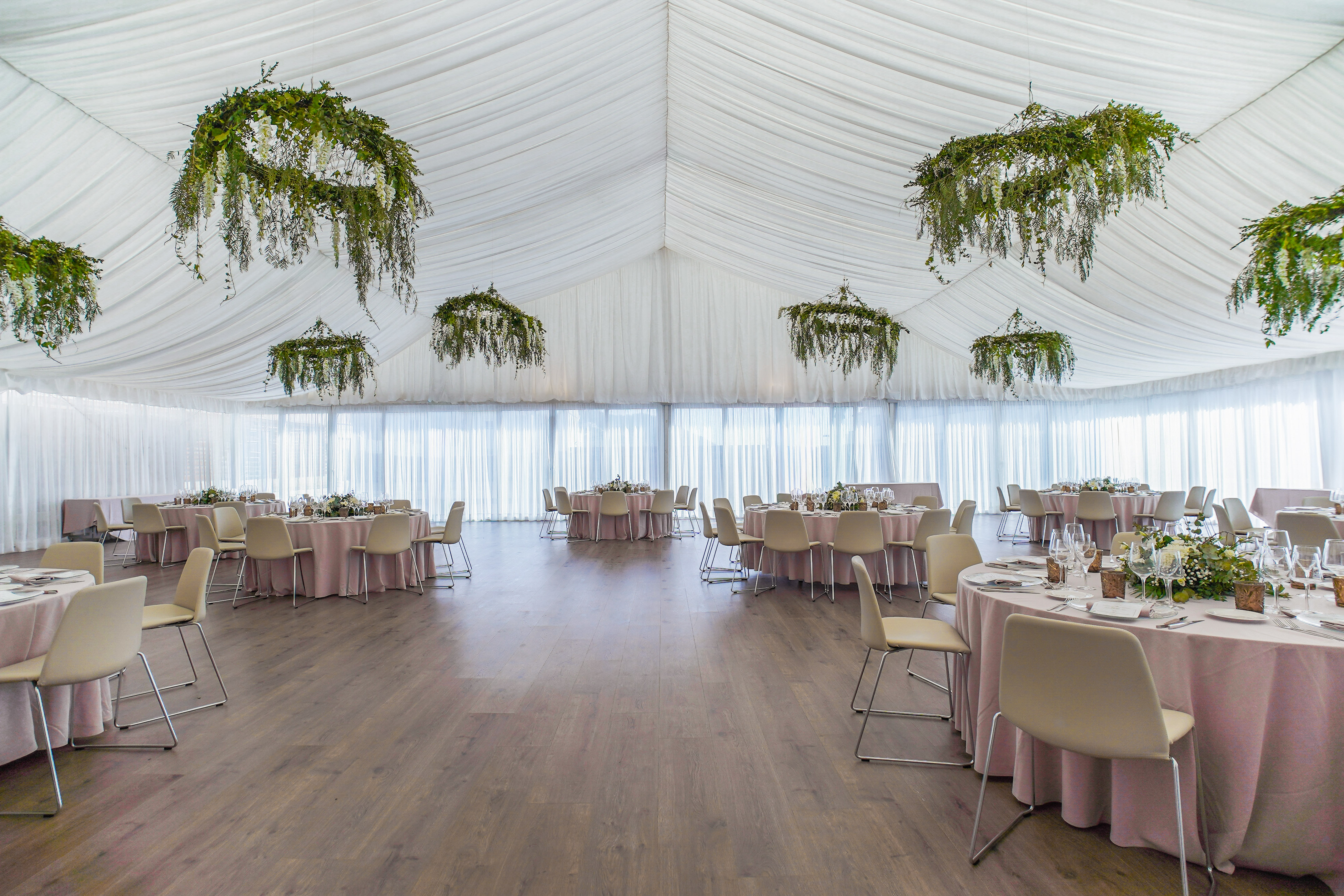 Marquee event space with banquet tables, white drapes, and greenery chandeliers.