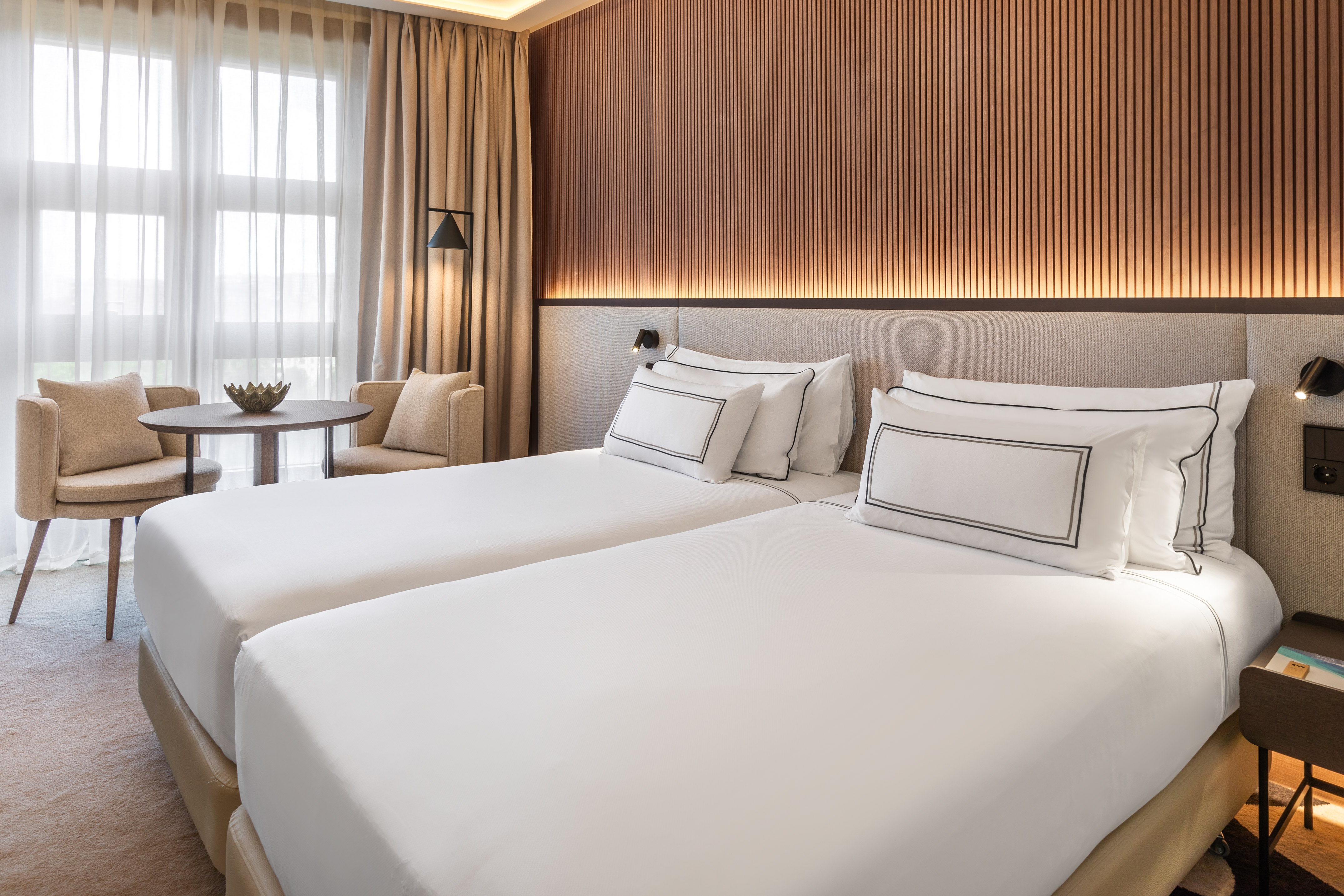 Modern hotel room with twin beds, wood-paneled wall, and window seating.