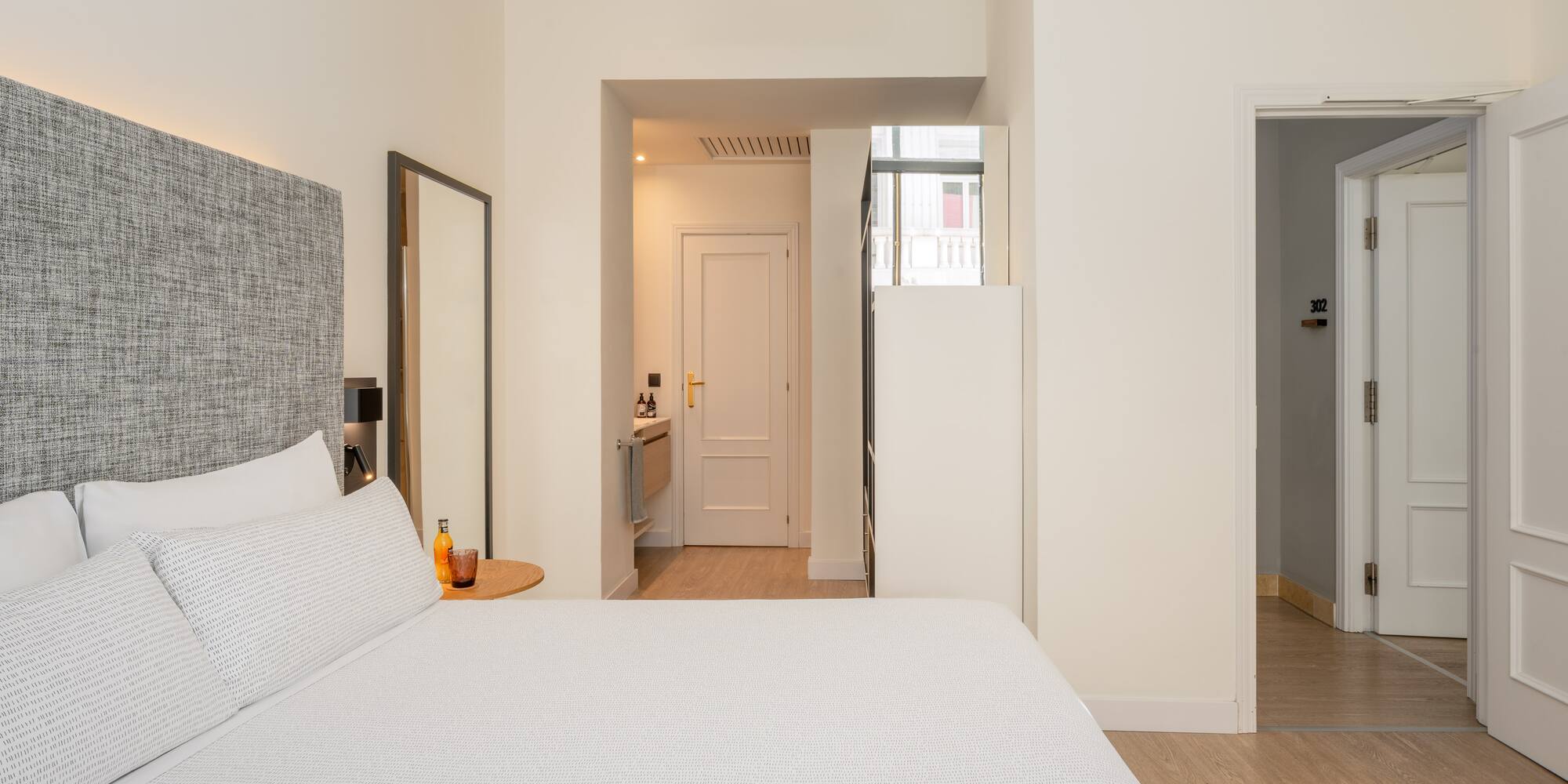 Modern hotel bedroom with white bed, grey headboard, and light wood floor.